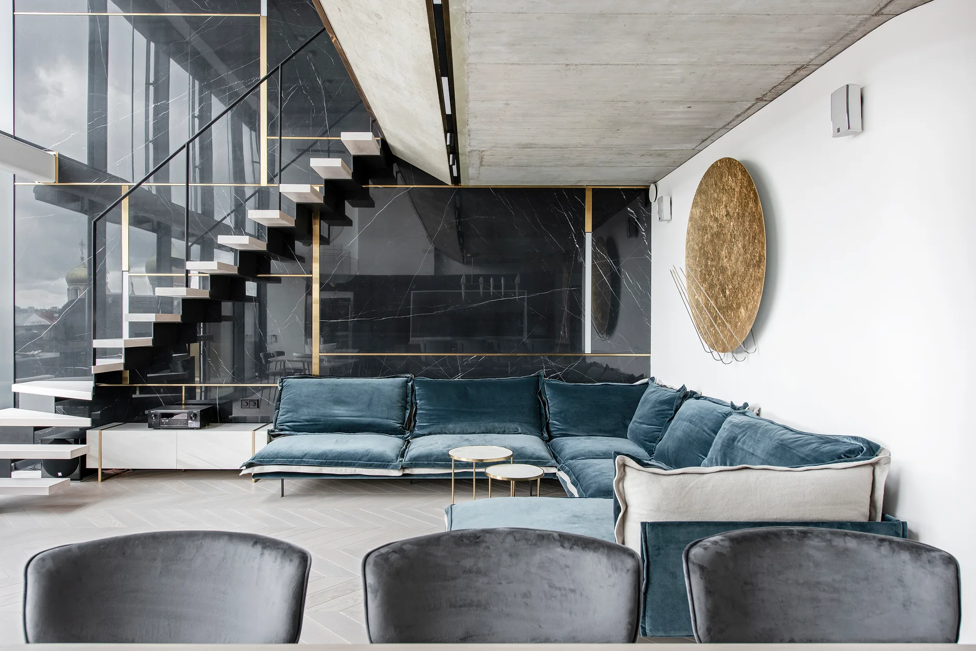 A modern living room features a teal velvet sectional sofa, a black marble wall with brass accents, and a floating staircase under a concrete ceiling.