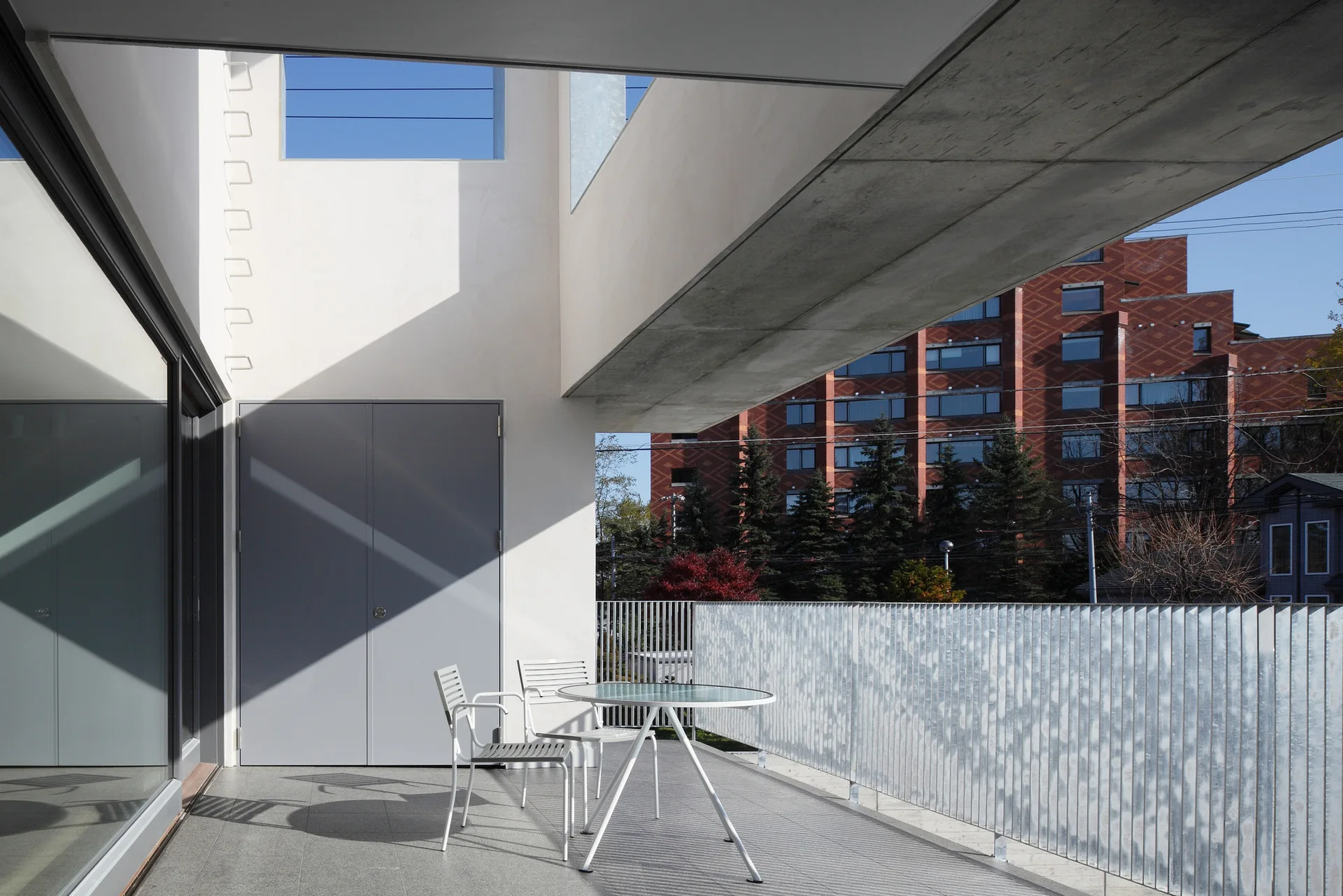 A modern minimalist balcony with a white metal bistro table and chairs on a grey tiled floor beneath a concrete beam.