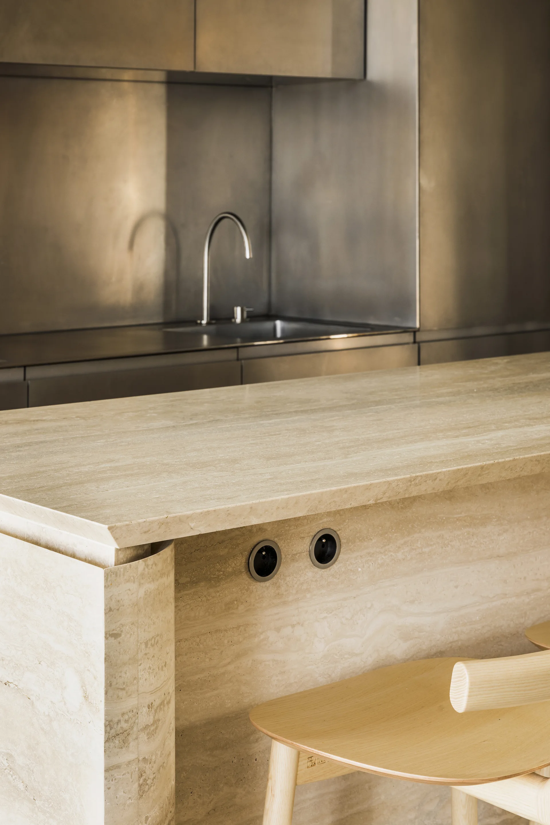 A minimalist kitchen features a beige travertine stone island with wooden stools next to brushed metal cabinetry with an integrated sink and faucet.
