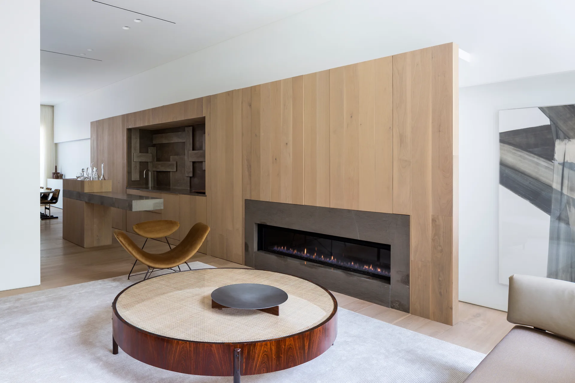 A modern living room features a vertical wood-paneled feature wall with a linear fireplace and integrated bar next to a round cane coffee table.