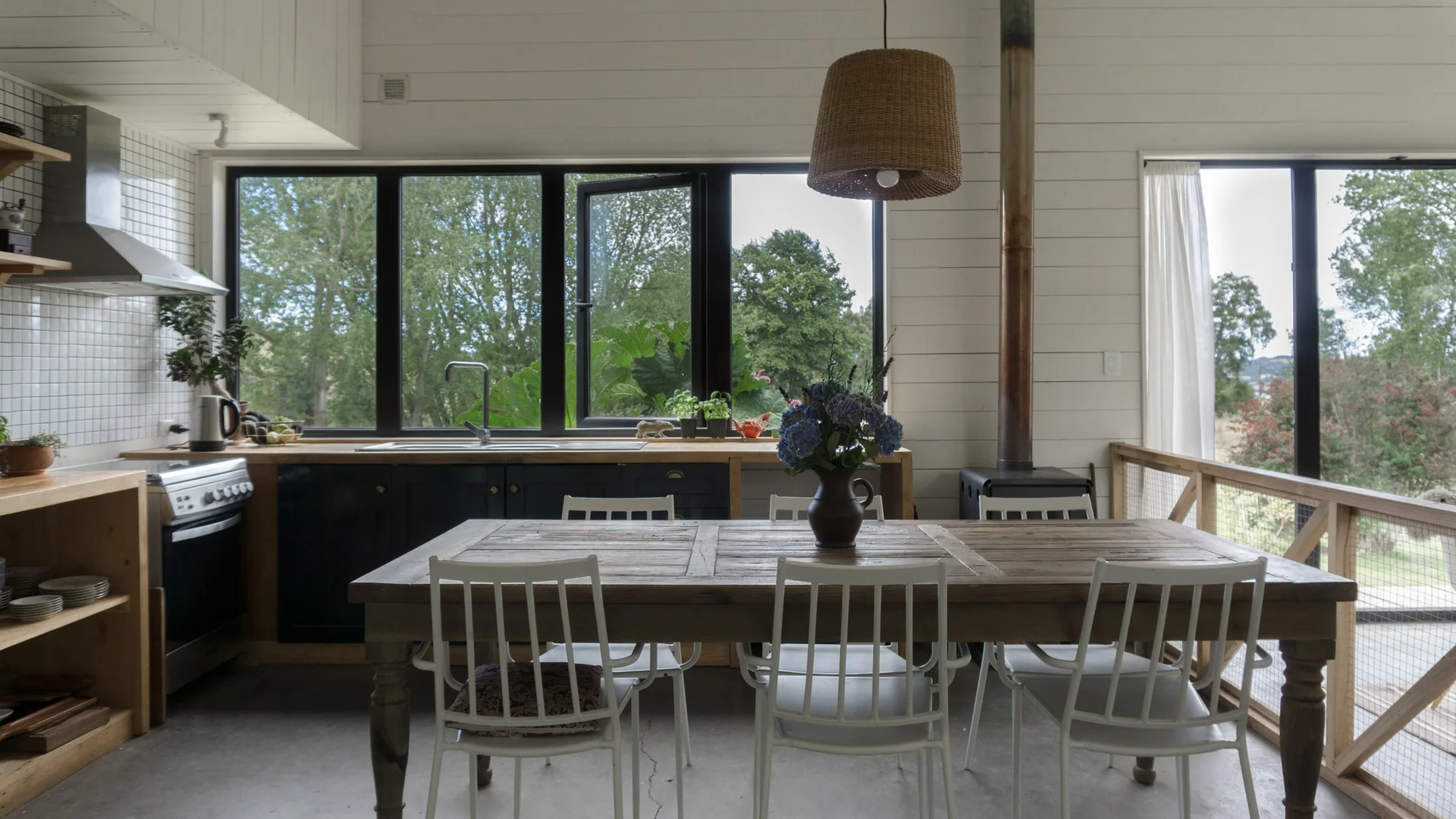 A rustic wood dining table with white chairs sits in a kitchen with white shiplap walls, dark cabinets, and large black-framed windows.