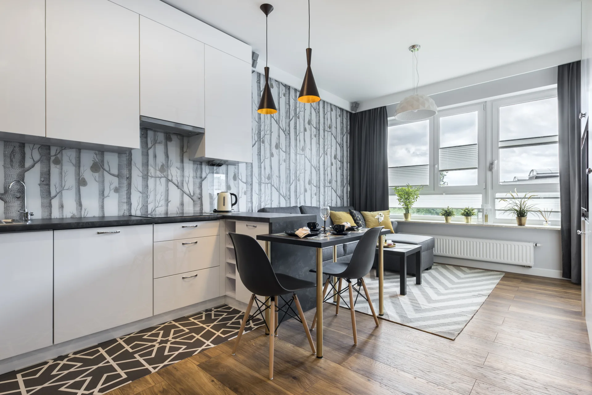A modern open-plan kitchen features white cabinetry, tree-patterned wallpaper, a small black dining set, and a grey sofa in the background.