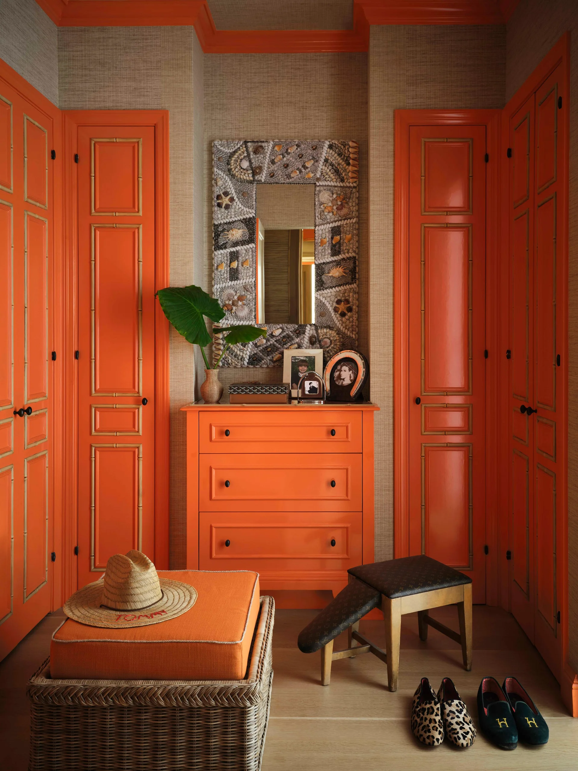 A dressing room features vibrant orange glossy cabinets, a central orange dresser, textured beige grasscloth wallpaper, and a shell-framed mirror.
