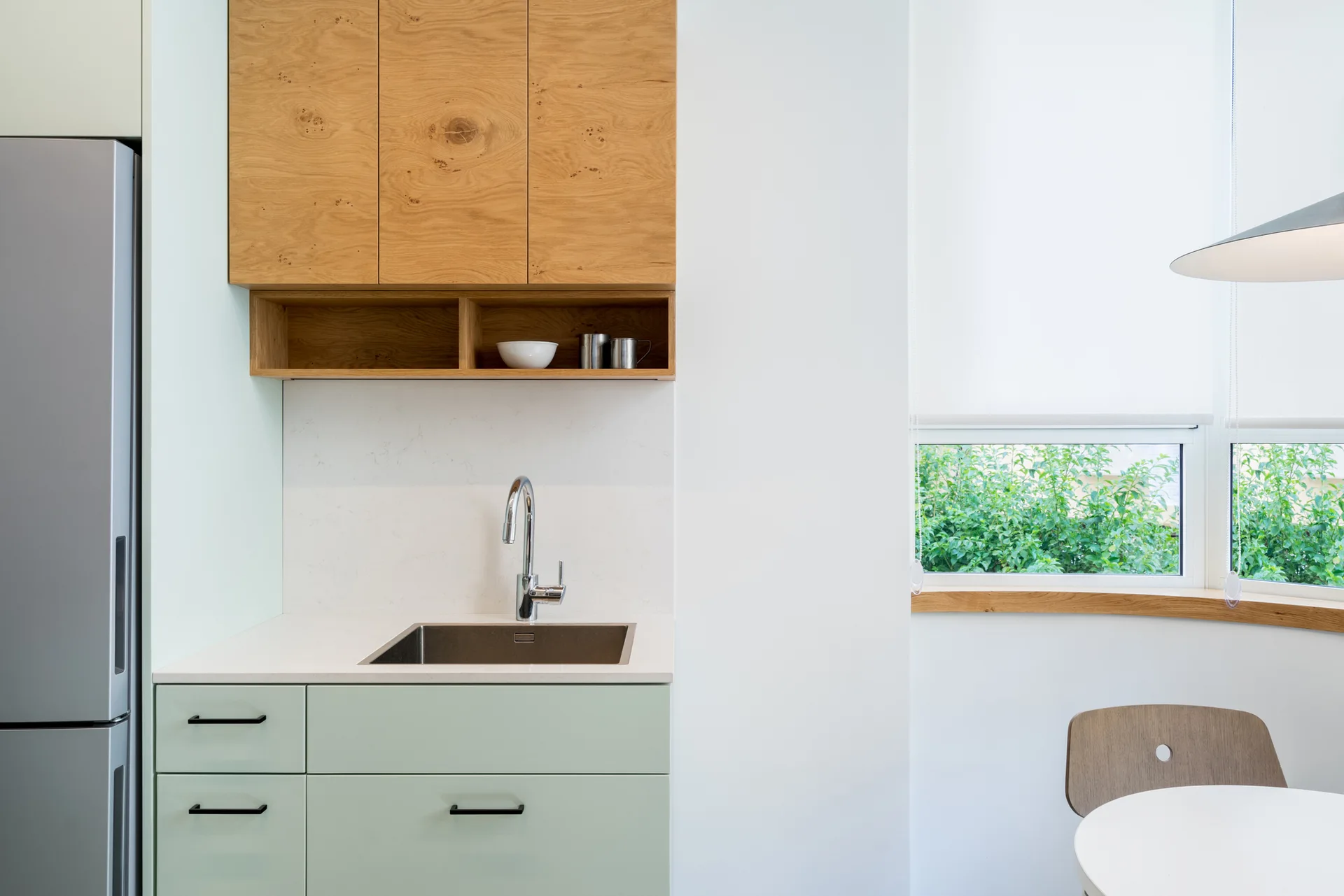 A kitchen corner features sage green lower cabinets, a stainless steel sink, natural wood upper cabinets, and a curved window nook on the right.