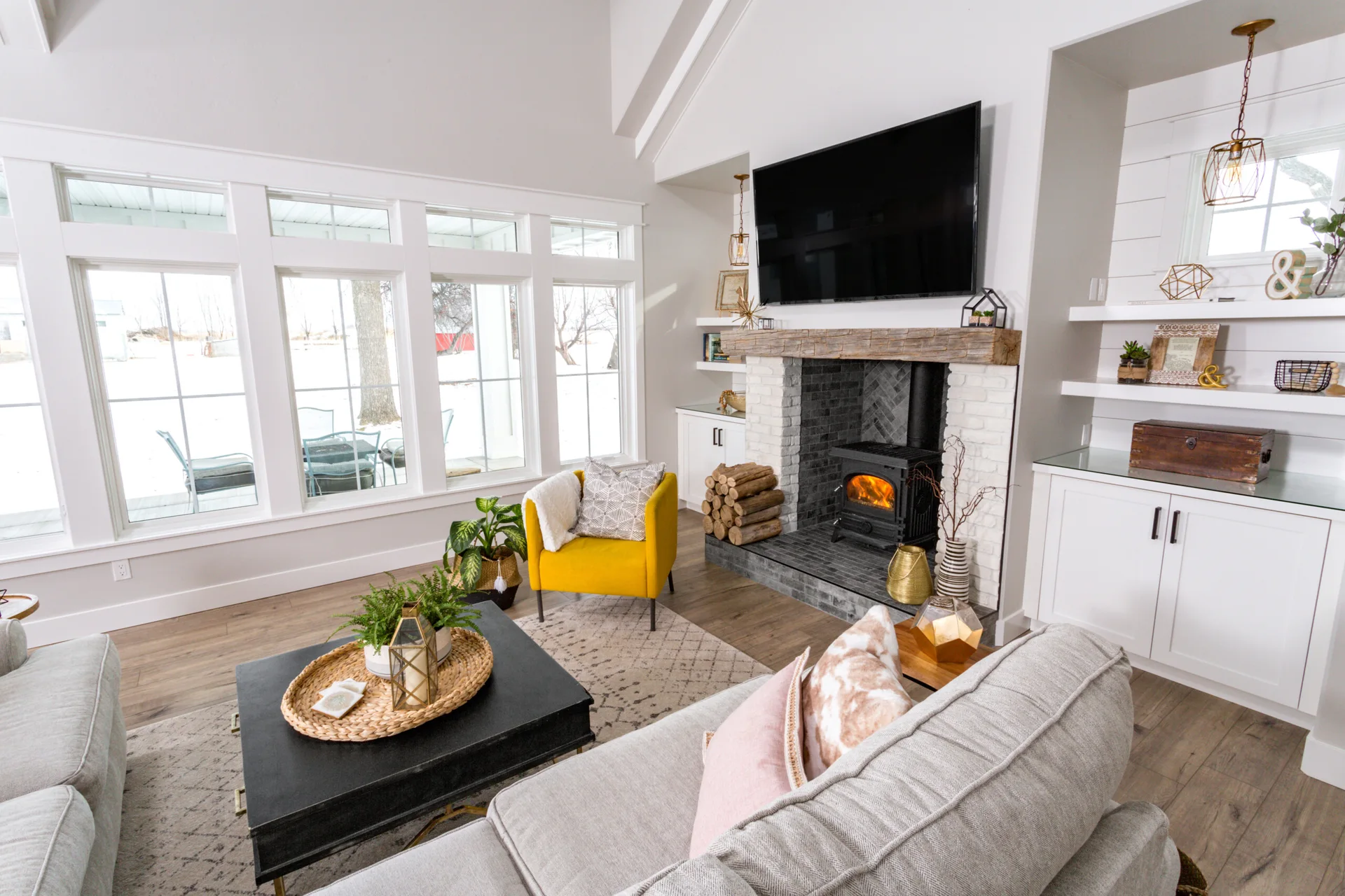 A bright living room features a vaulted ceiling, a white brick fireplace with a wood mantel, a grey sofa, and a yellow armchair.