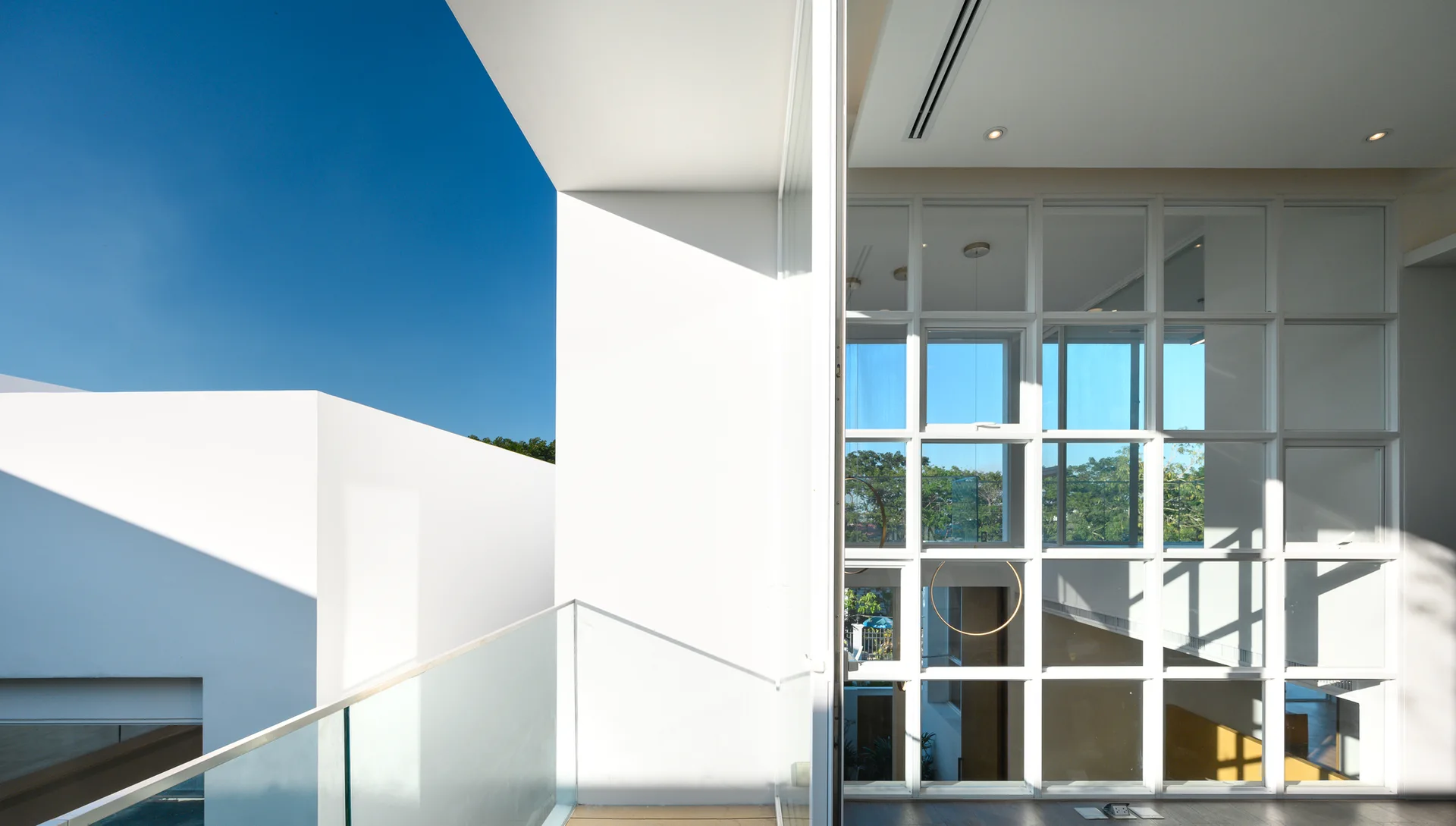 A modern white interior features a large grid-patterned window wall next to an outdoor balcony with a glass railing under a bright blue sky.