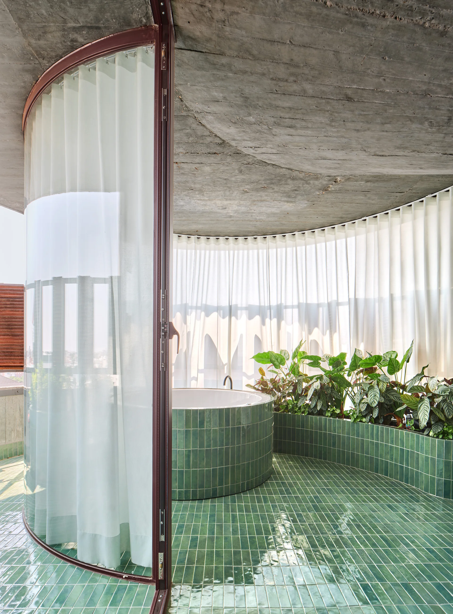 A curved bathroom featuring glossy green tiled floors and walls, a circular bathtub, a built-in planter with green foliage, and a raw concrete ceiling.