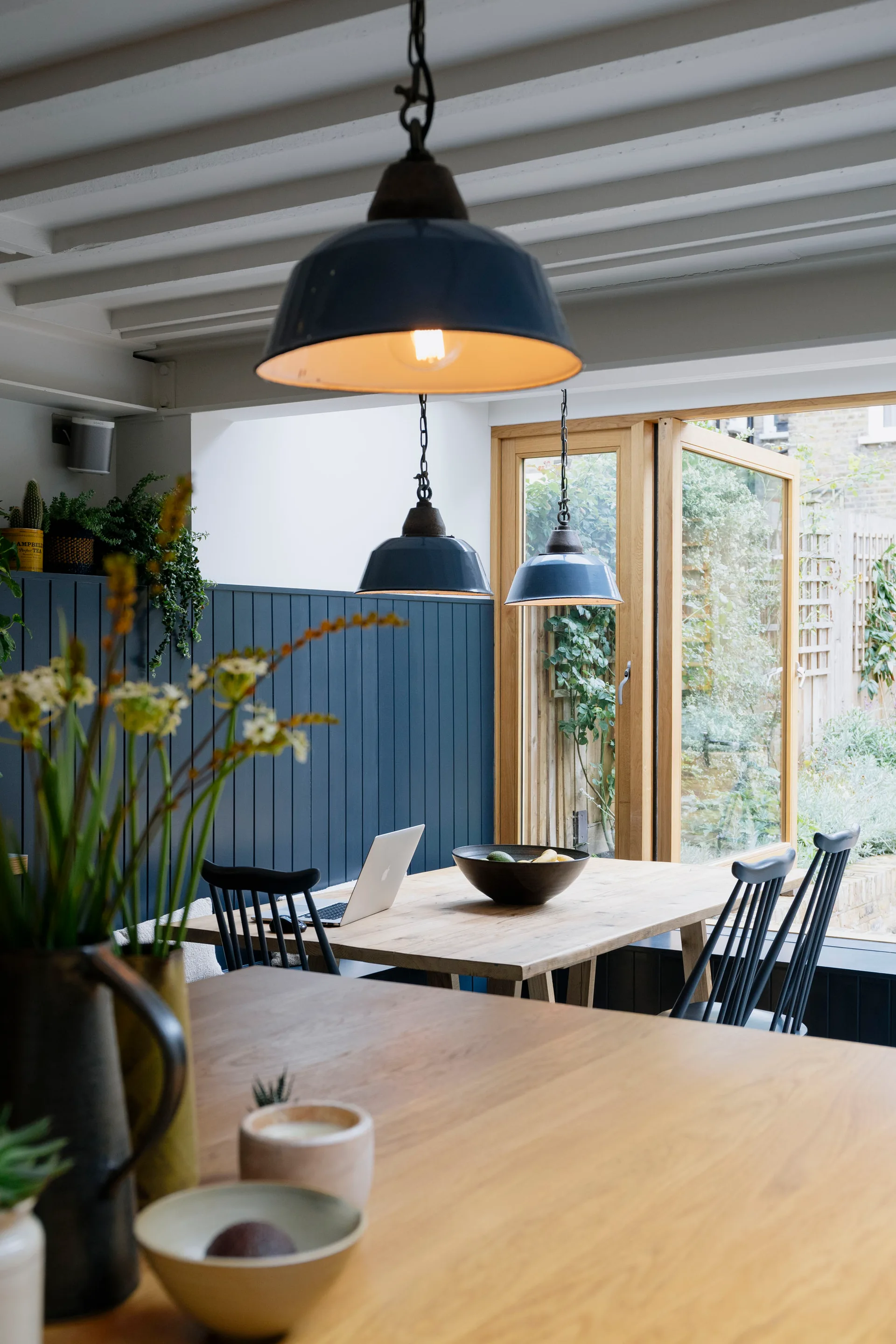 A light wood dining table with black spindle chairs sits before a navy blue paneled wall and large wood-framed windows overlooking a green garden.