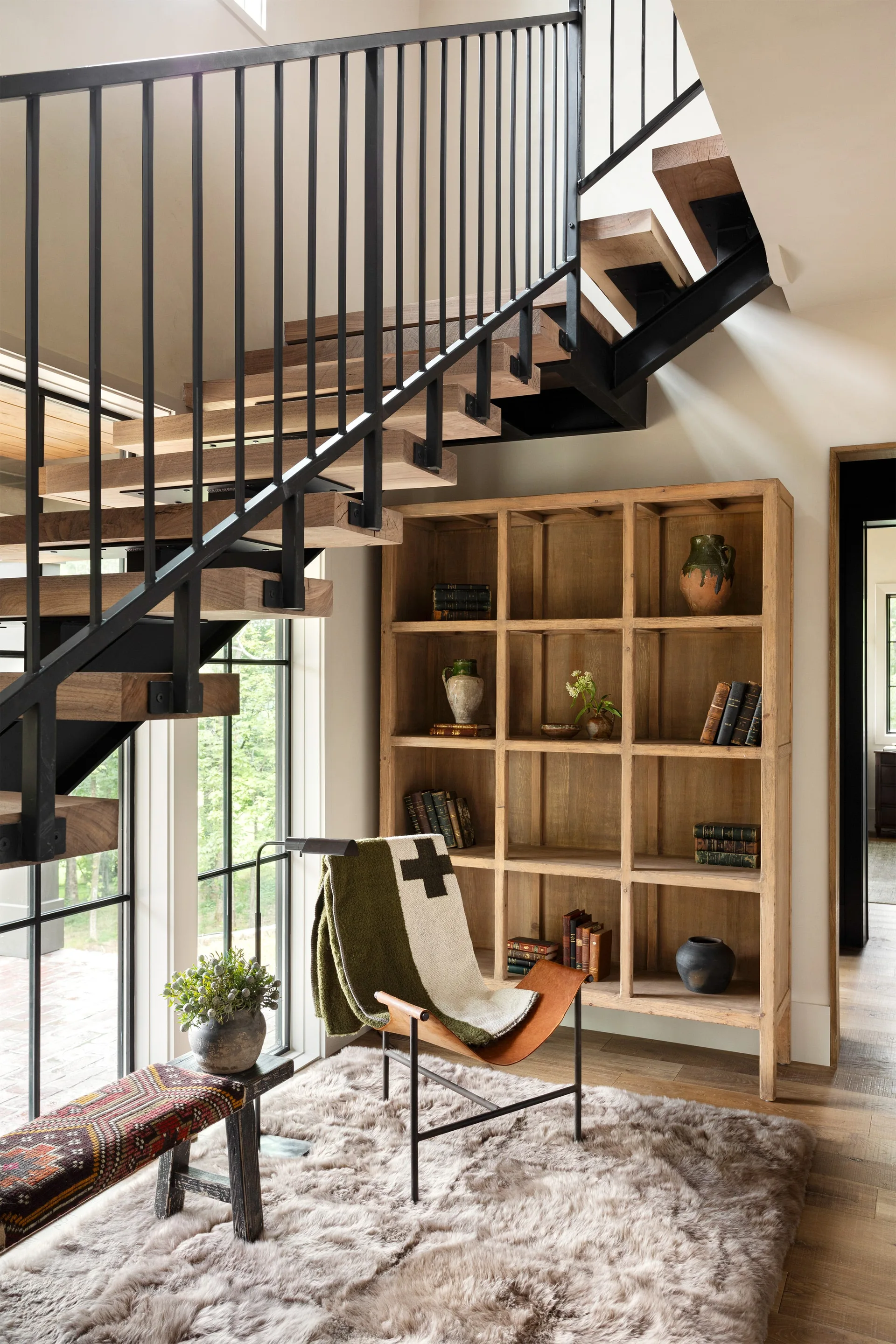 A cozy reading nook with a leather sling chair and a wooden grid bookshelf sits under a modern open-tread staircase with black metal railings.