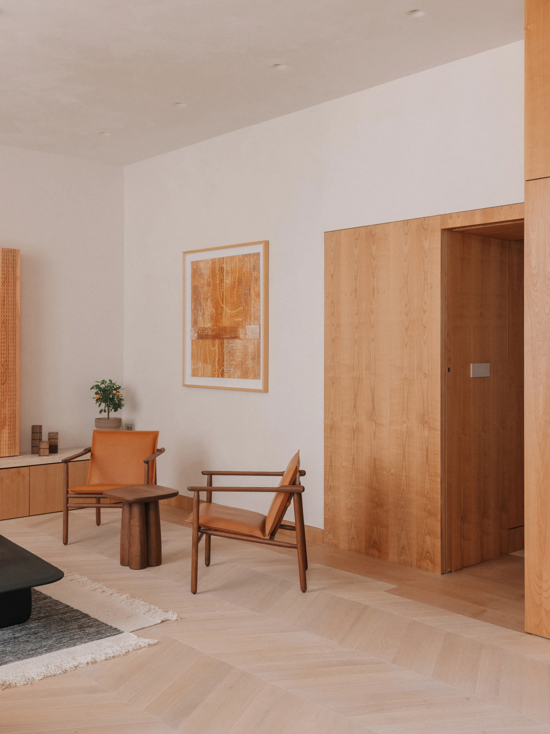 Two tan leather armchairs and a small wooden side table sit on a light wood herringbone floor next to a wood-paneled doorway.