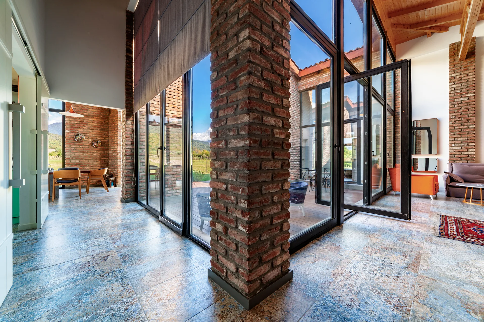 A spacious open-concept room features prominent exposed brick columns, patterned blue-toned floor tiles, and tall black-framed glass walls.