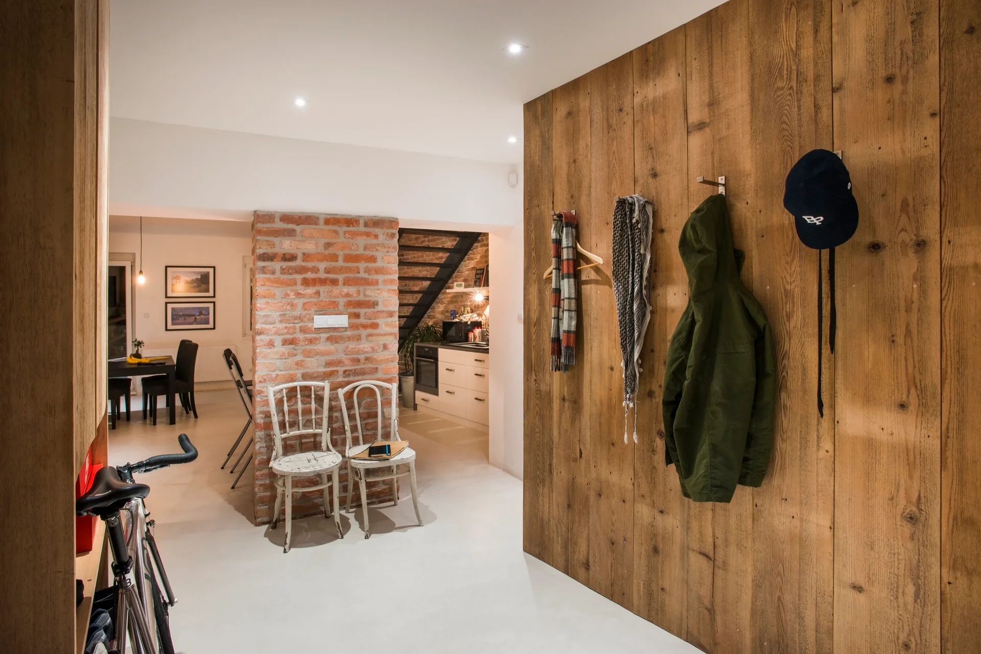 An entryway featuring a wood-paneled accent wall with coat hooks, two distressed white chairs against an exposed brick pillar, and smooth flooring.