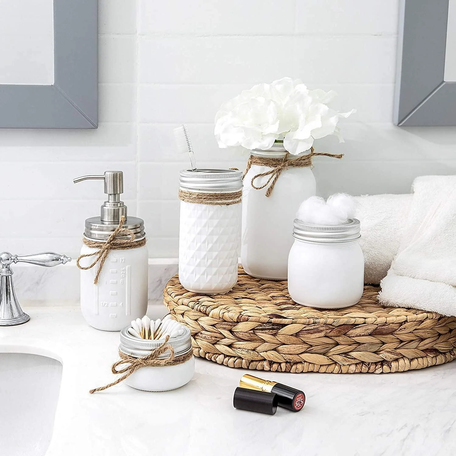 White painted mason jar bathroom accessories and a woven natural fiber tray sit on a marble vanity top against a subway tile backsplash.