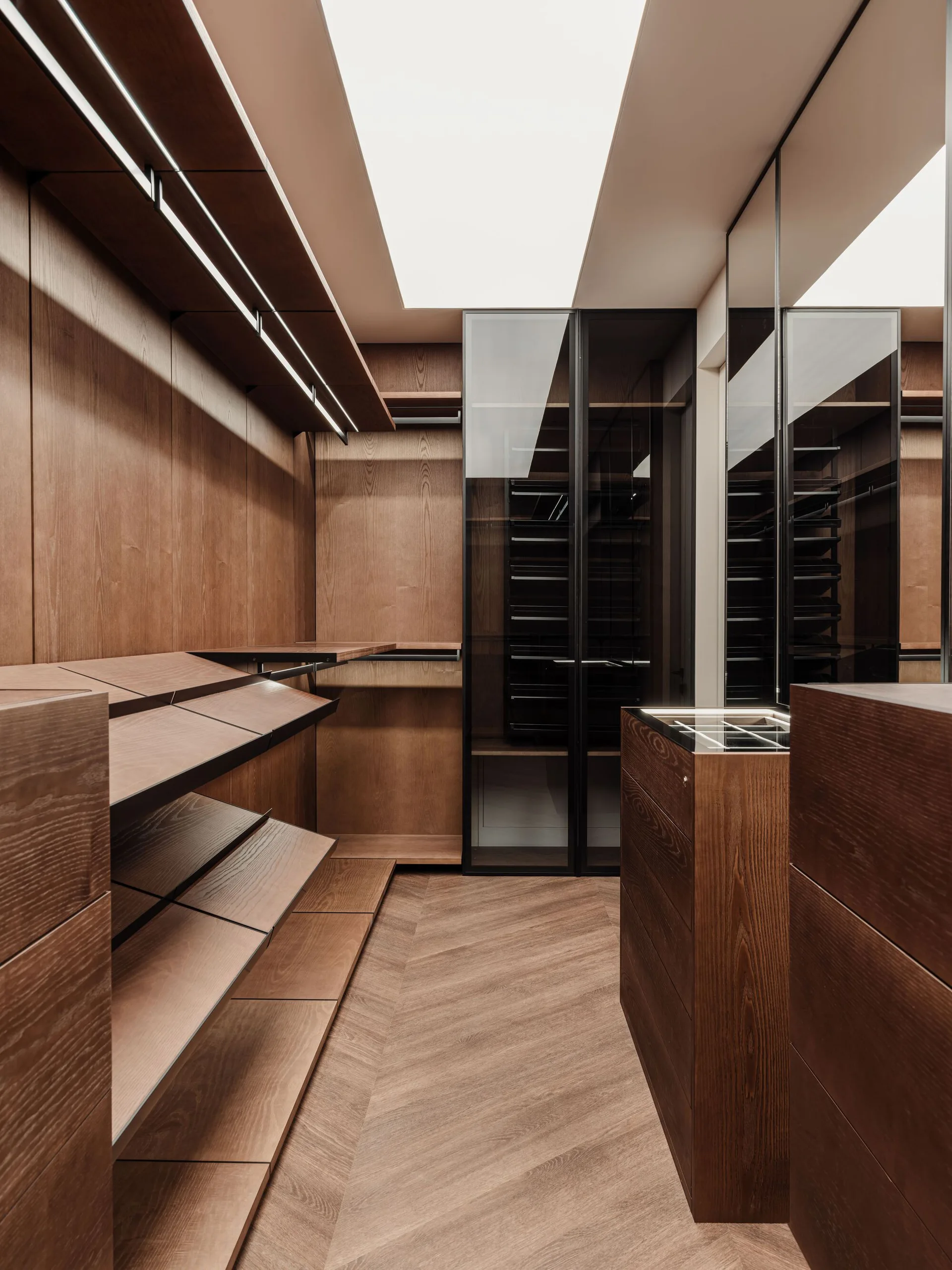 A modern walk-in closet features warm wood cabinetry, a large ceiling skylight, chevron-patterned wood floors, and integrated shelf lighting.