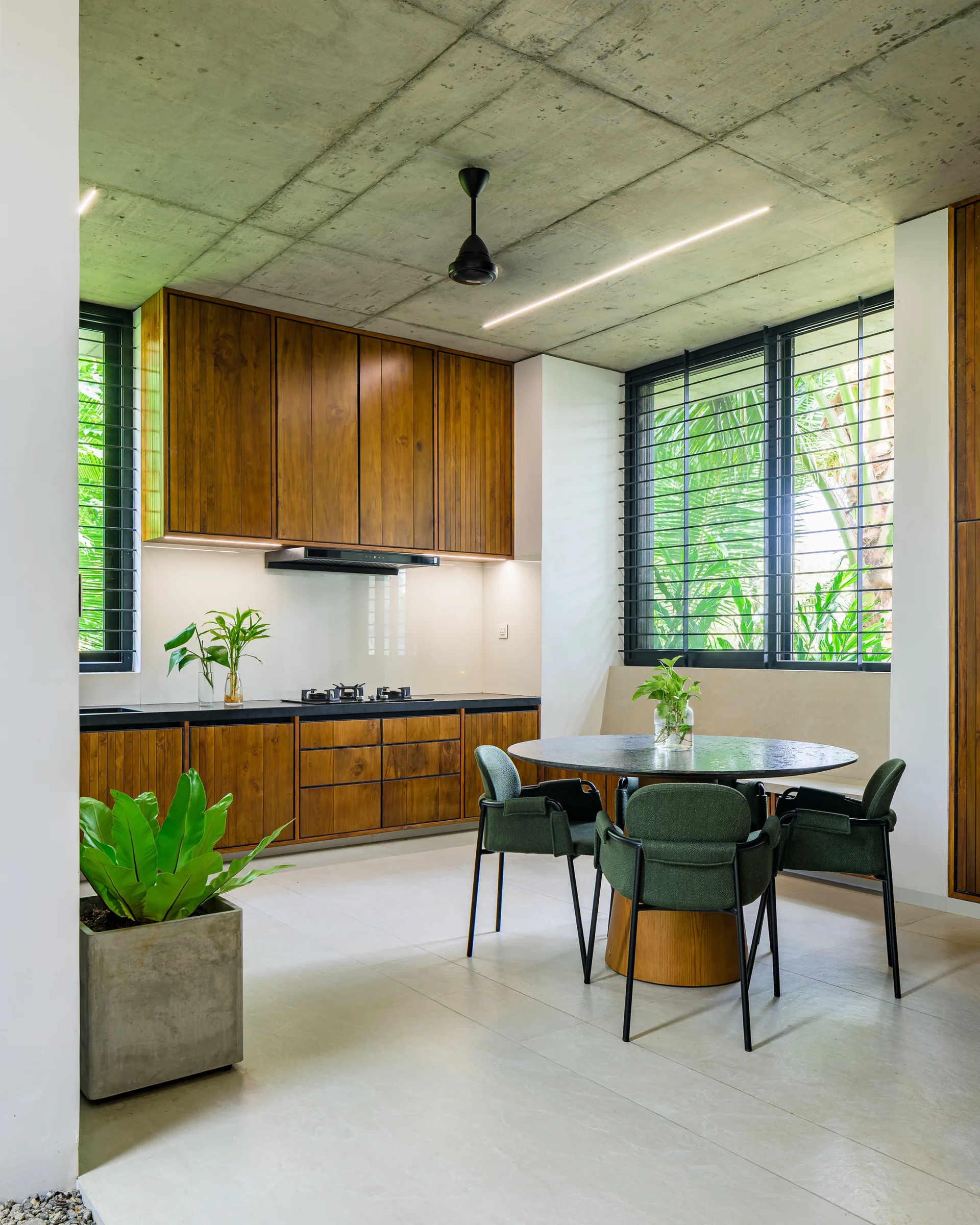 A modern kitchen and dining space with an exposed concrete ceiling, warm wood cabinetry, a circular dining table, and green upholstered chairs.