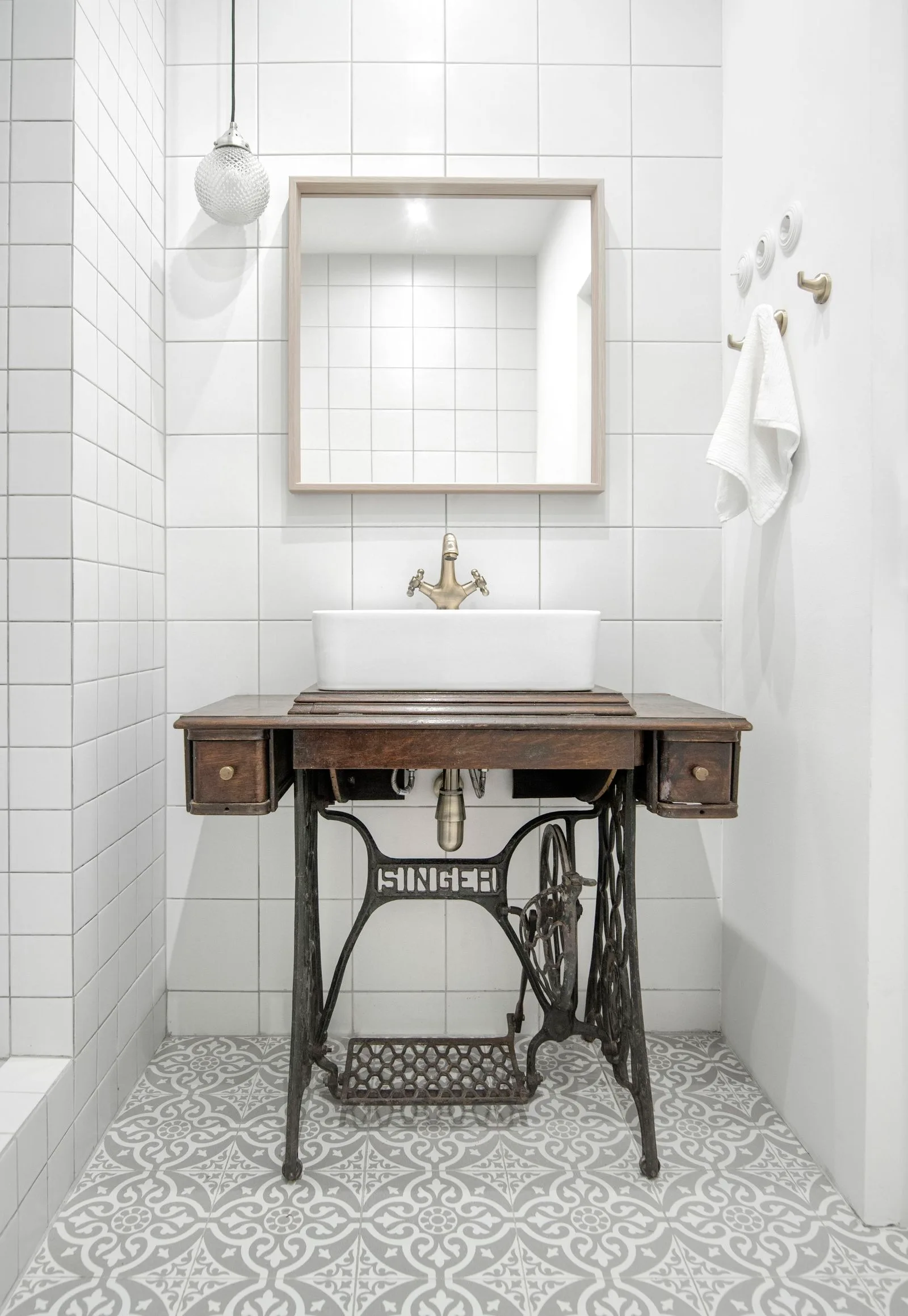 A white vessel sink sits on a repurposed vintage sewing machine base in a powder room with white grid wall tiles and patterned floors.