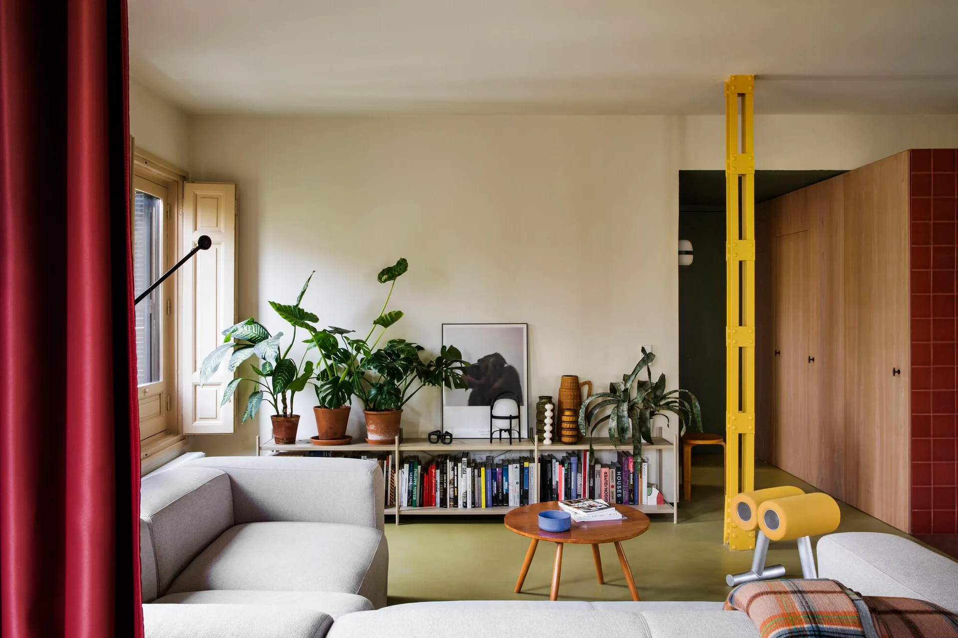 An eclectic living room featuring a grey sectional sofa, a low wooden bookshelf with potted plants, and a prominent bright yellow structural column.
