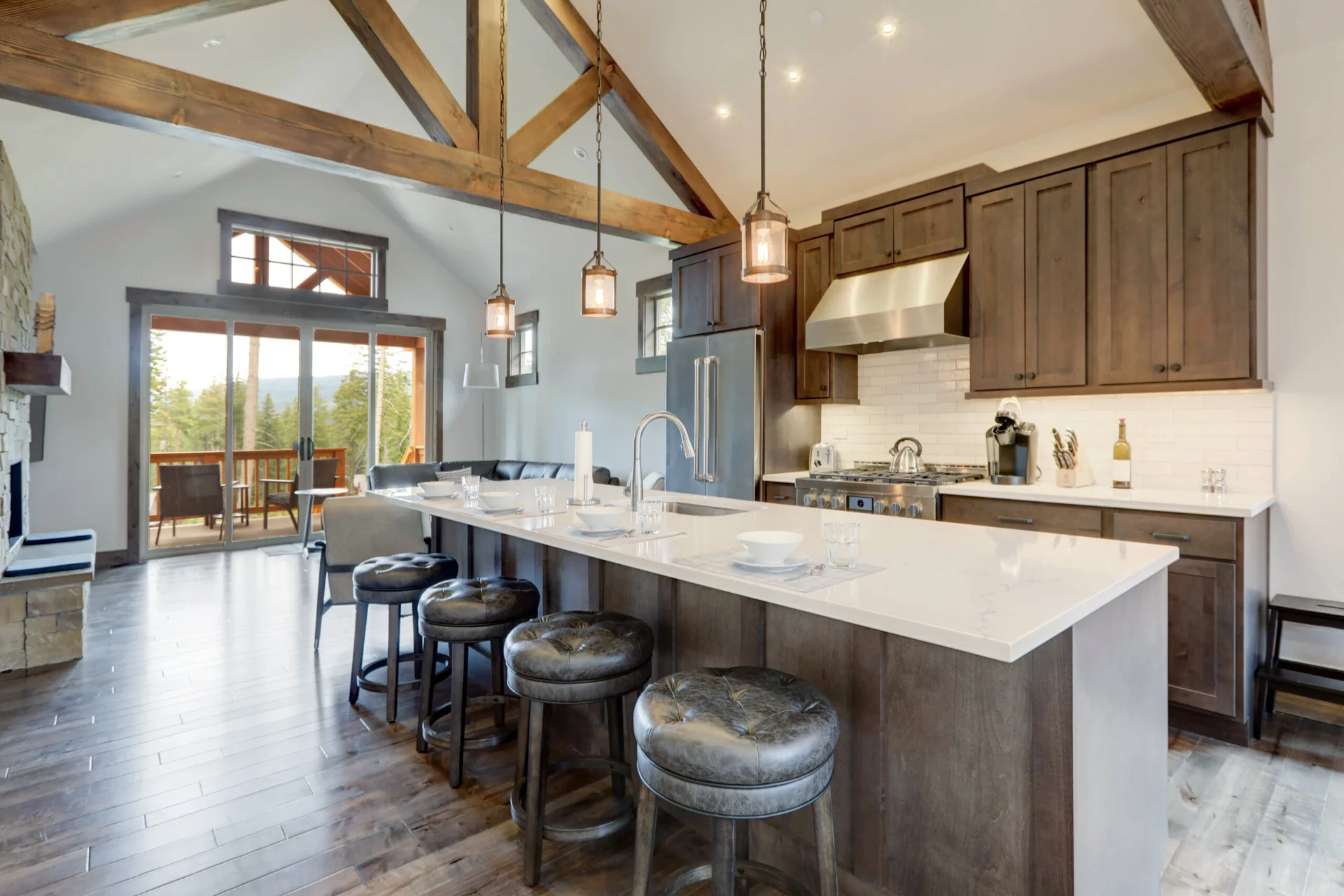 A kitchen featuring a white island with four dark tufted stools, dark wood cabinets, a subway tile backsplash, and exposed wooden ceiling beams.
