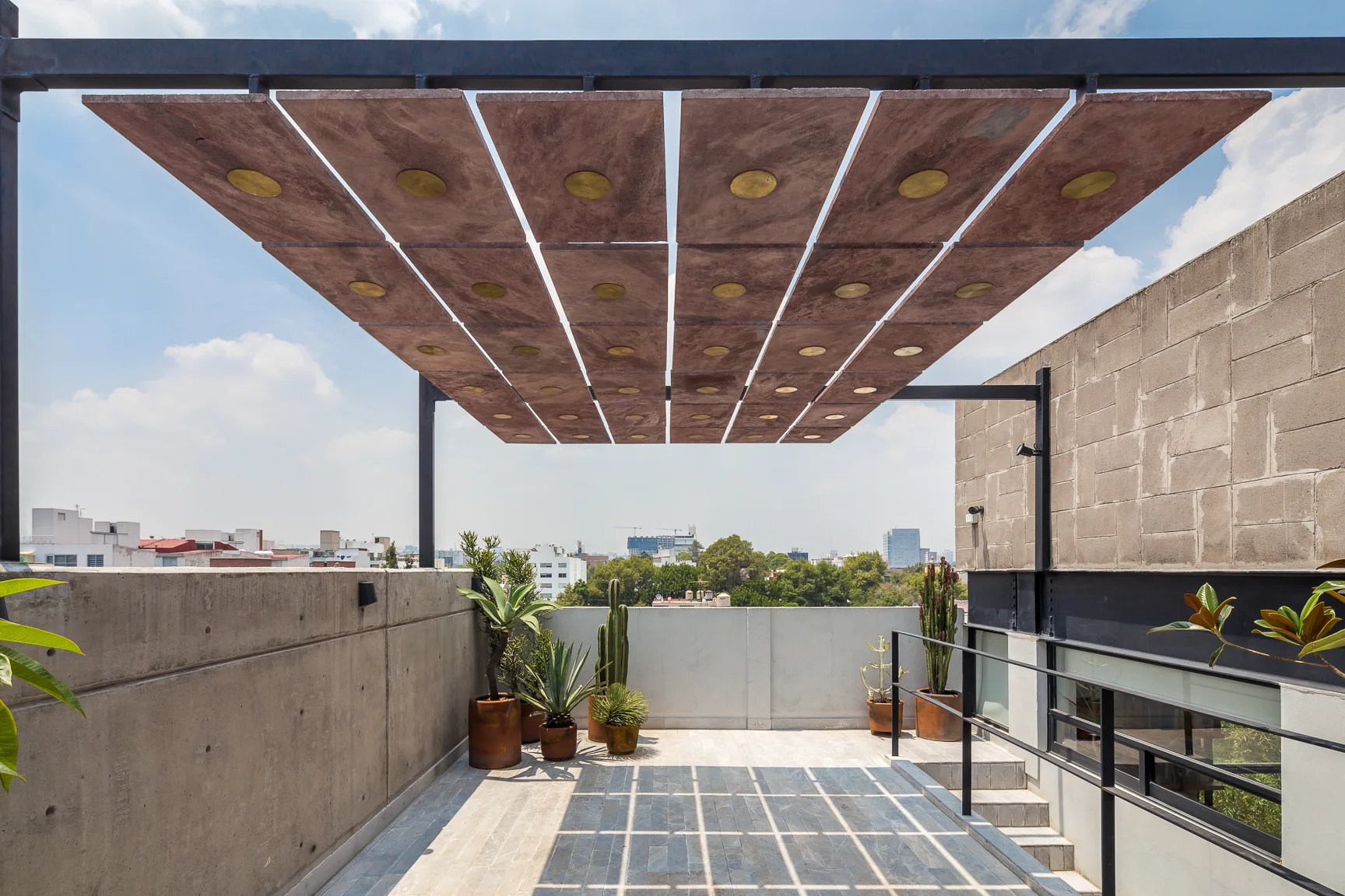 A modern rooftop terrace features a terracotta panel shade canopy supported by a black metal frame, concrete walls, and potted cacti.