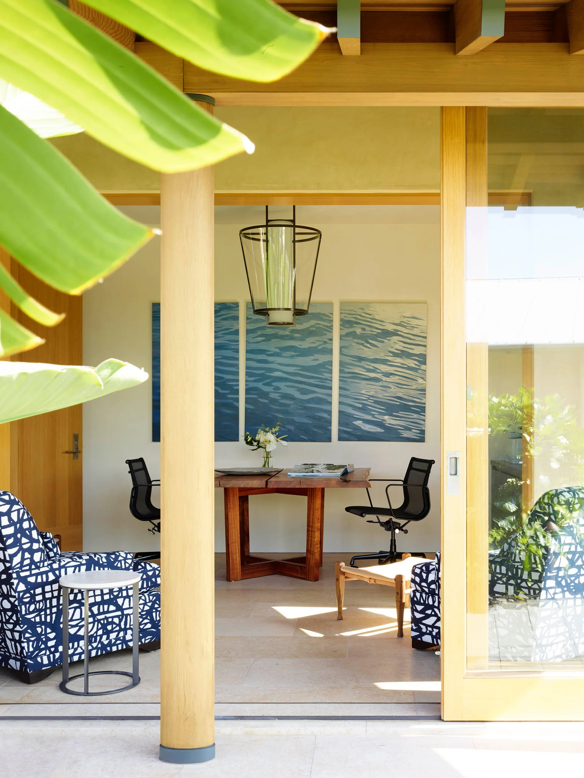 A sunlit home office featuring a wood desk and black mesh chairs, viewed through a sliding glass door beneath a blue triptych artwork.