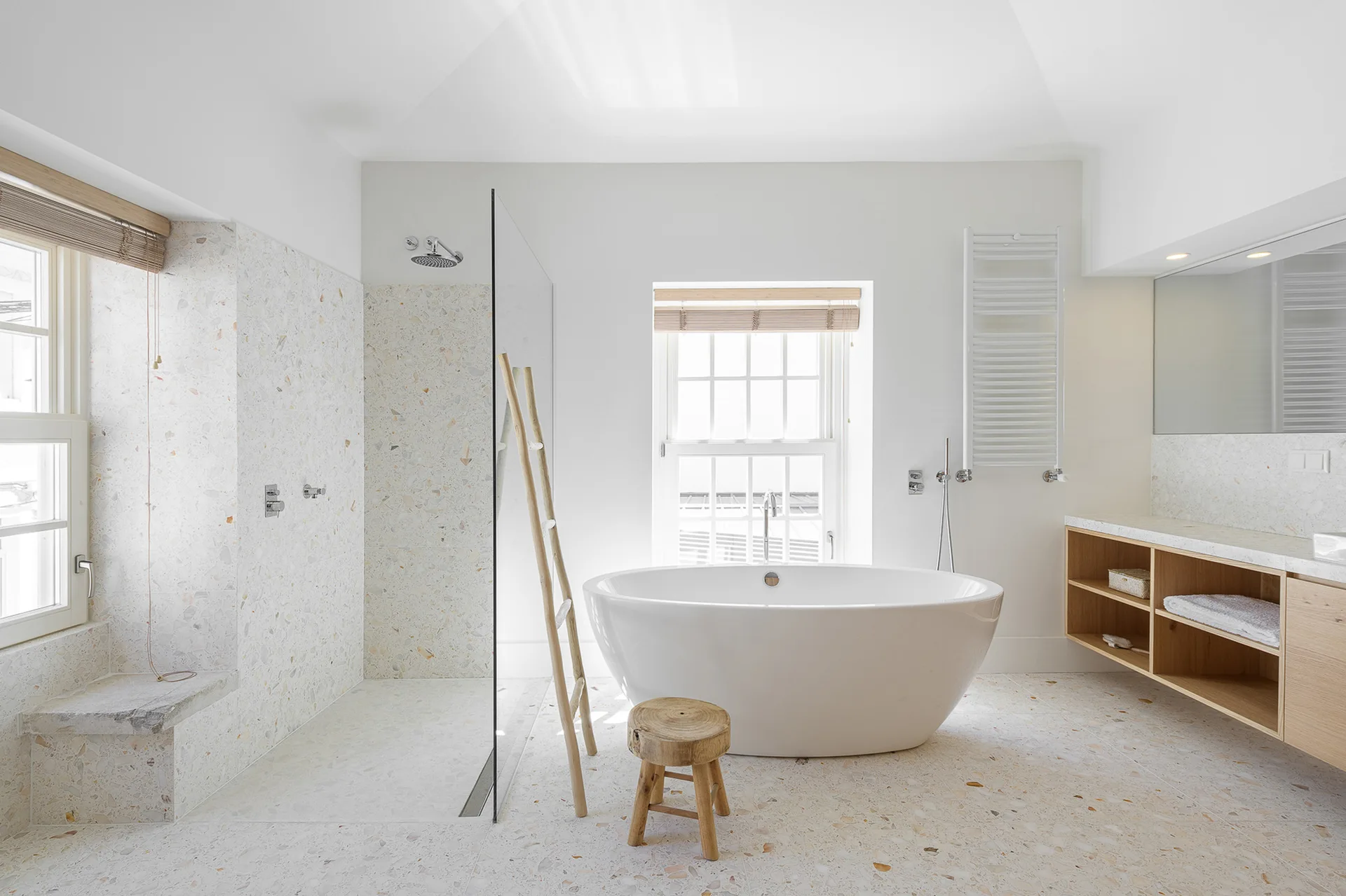 A bright bathroom features a white freestanding bathtub, a walk-in shower with speckled terrazzo walls, and a light wood floating vanity.