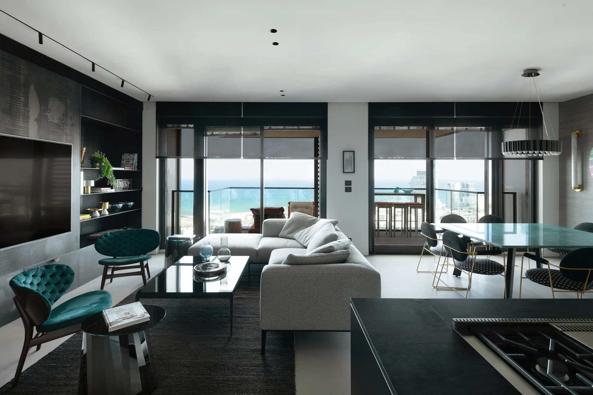 A modern open-concept living room features a grey sectional sofa, teal tufted accent chairs, and floor-to-ceiling windows overlooking the ocean.