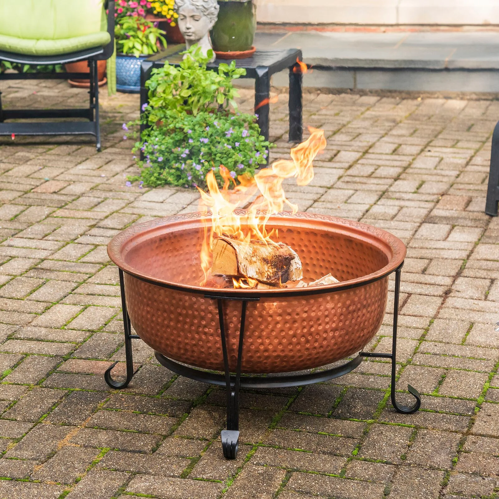 A hammered copper fire pit with burning wood logs sits on a stone paver patio next to green plants.