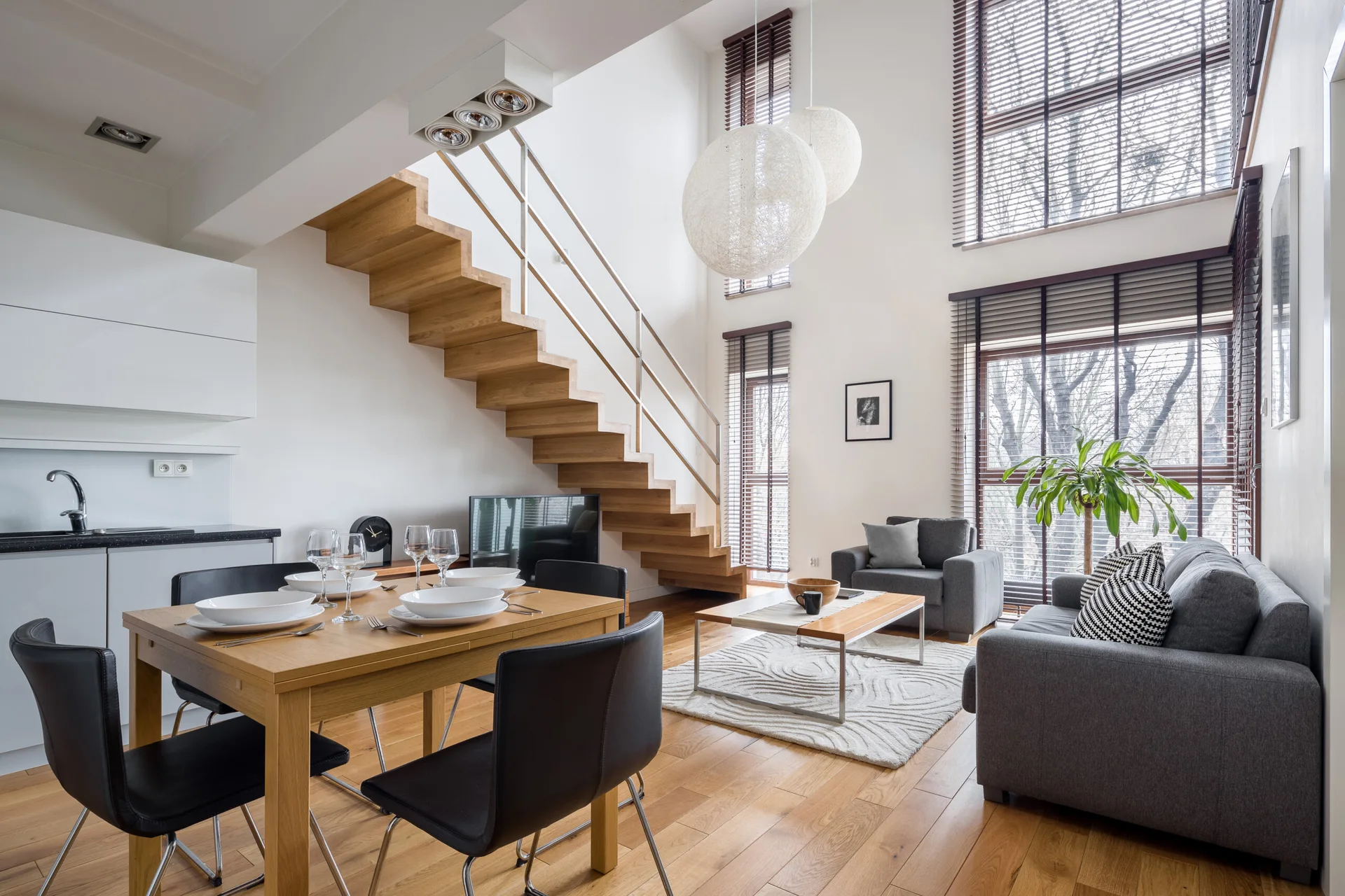 A bright open-plan living space featuring a floating wooden staircase, a gray sofa, a wood dining table, and large double-height windows.