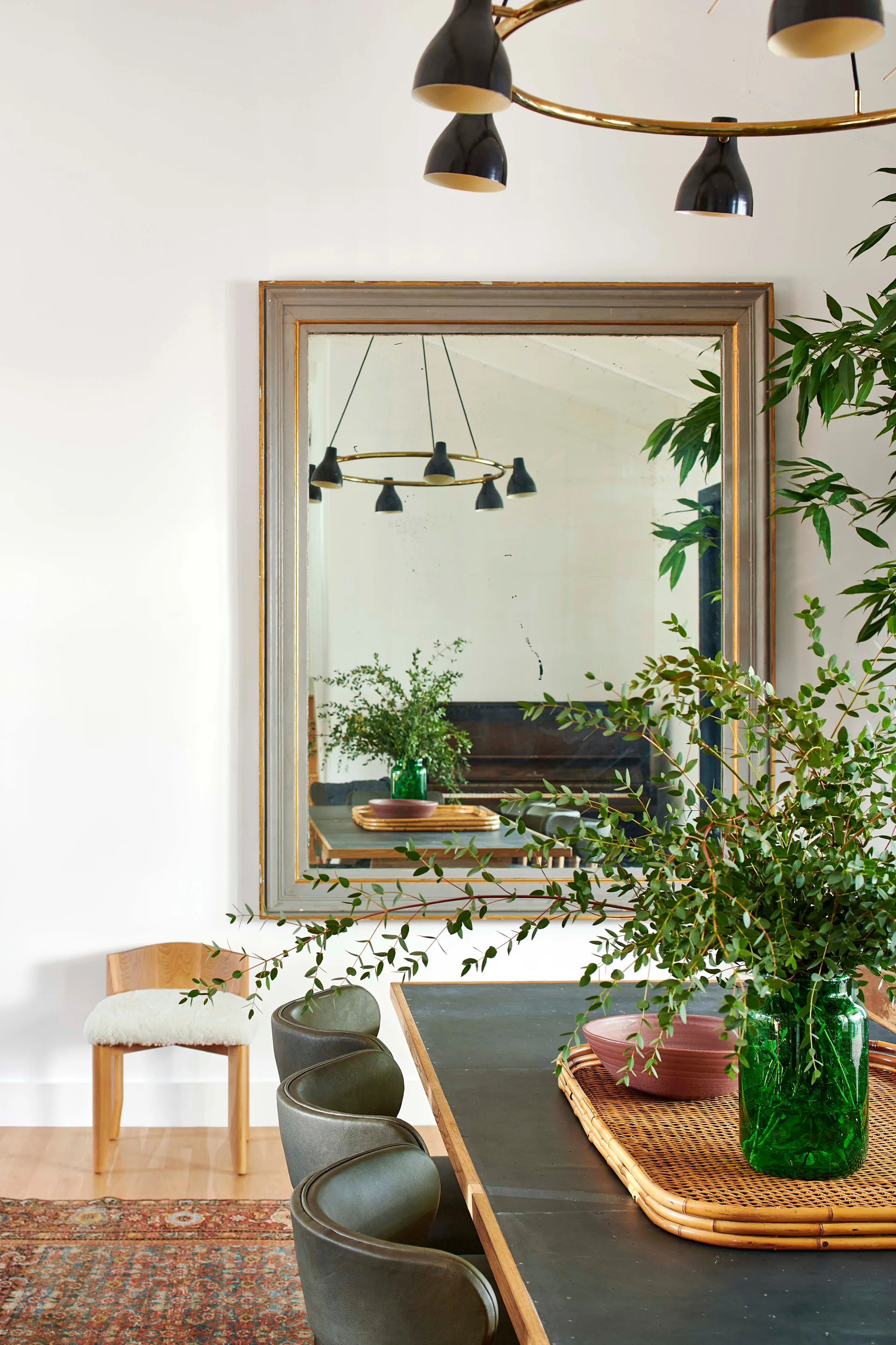 Olive green leather chairs surround a dark dining table beneath a brass circular chandelier, with a large framed mirror hanging on the white wall.