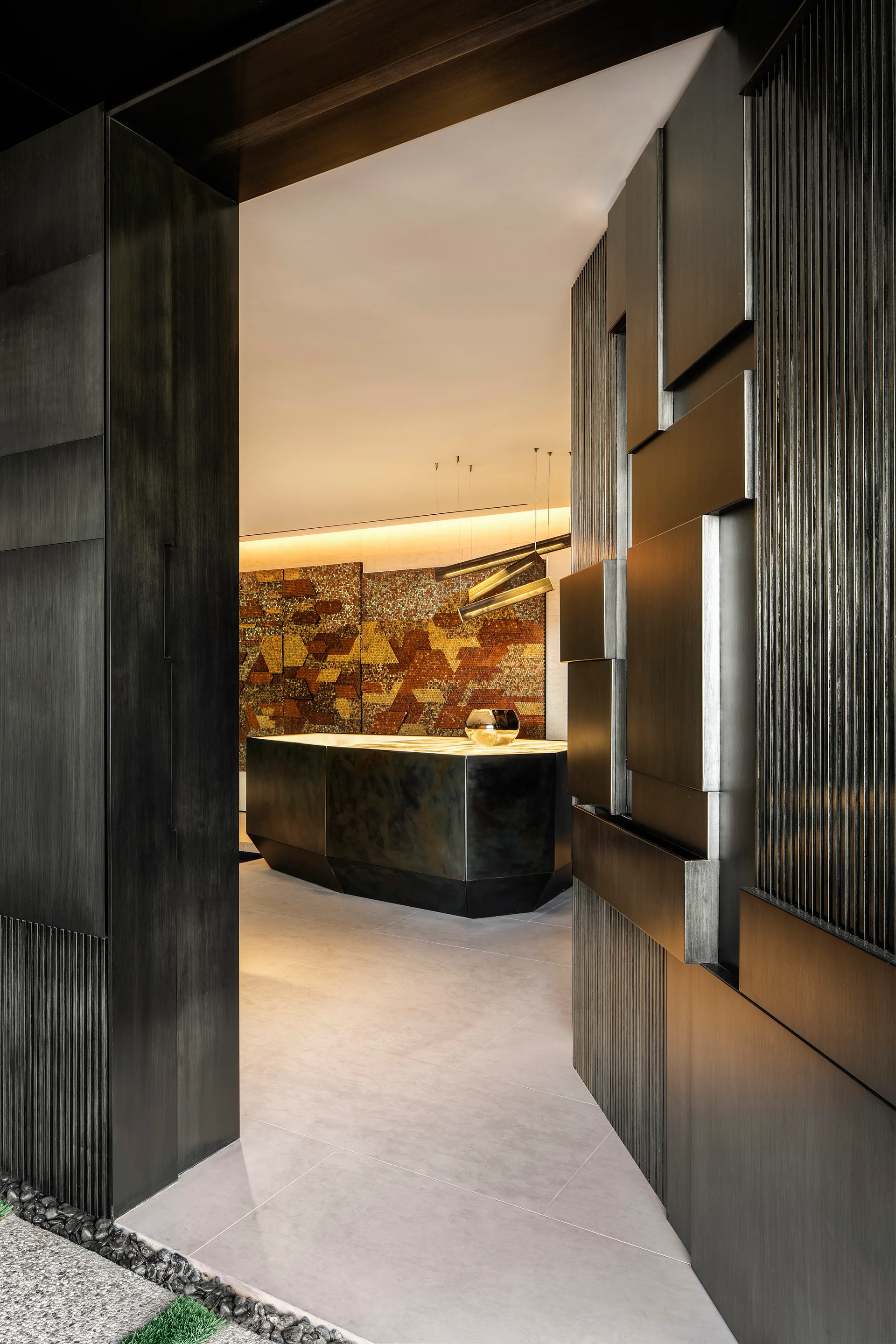 A modern entryway features a sculptural dark metal desk beneath linear pendant lights and a textured geometric wood wall panel in the foreground.