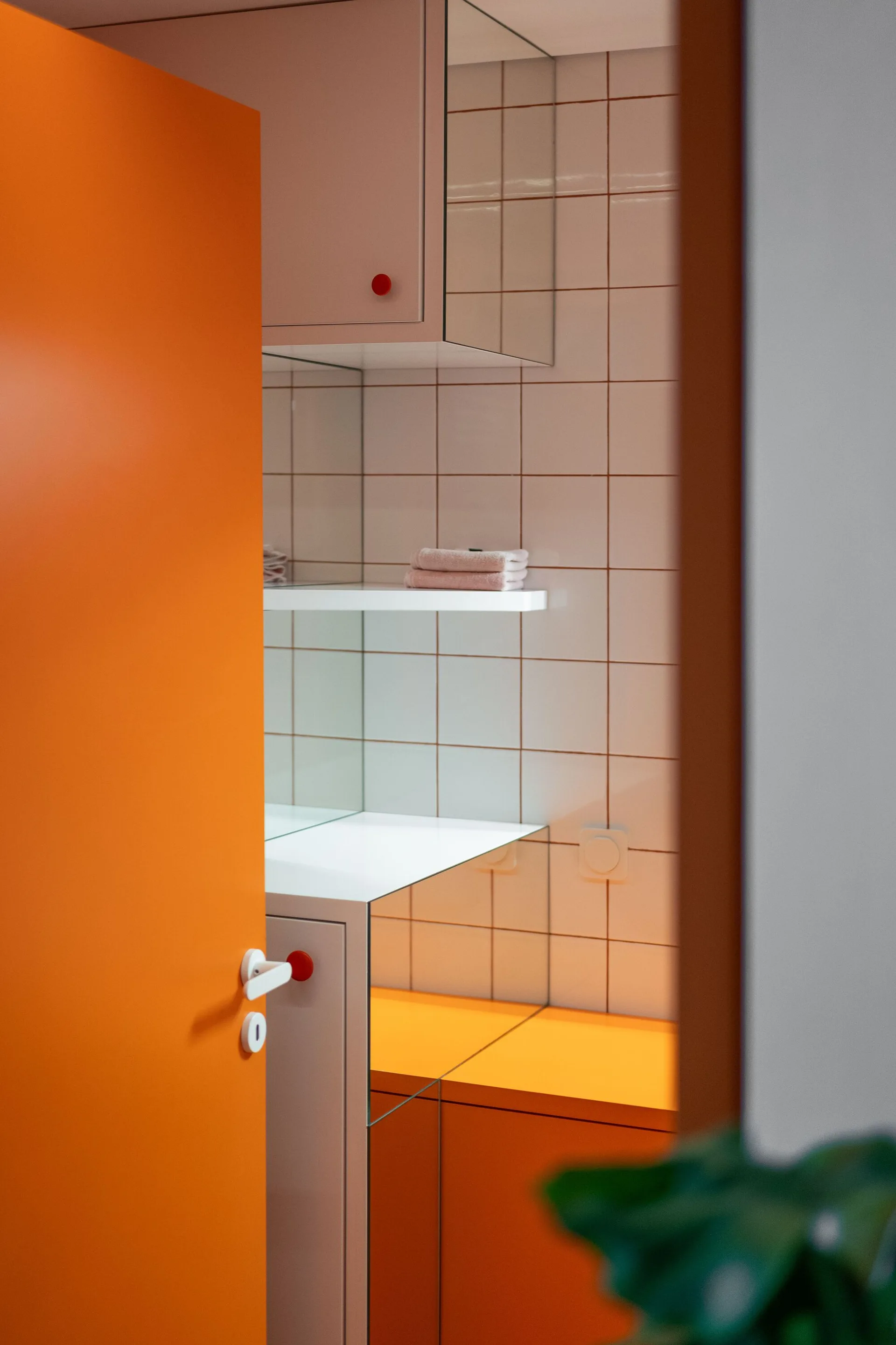 A view through a bright orange door into a bathroom with white grid wall tiles, a white floating shelf, and a mirrored vanity cabinet.
