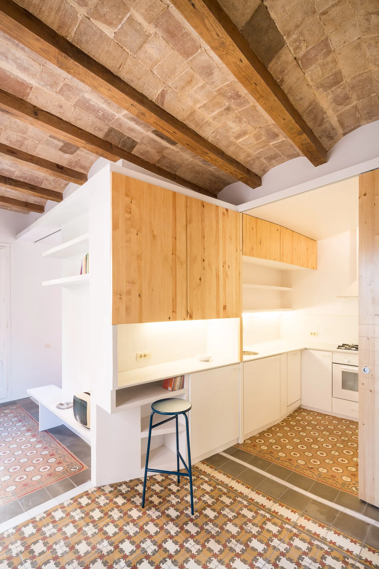A modern kitchen with a vaulted brick ceiling, wood and white cabinets, and patterned floor tiles next to a blue bar stool.