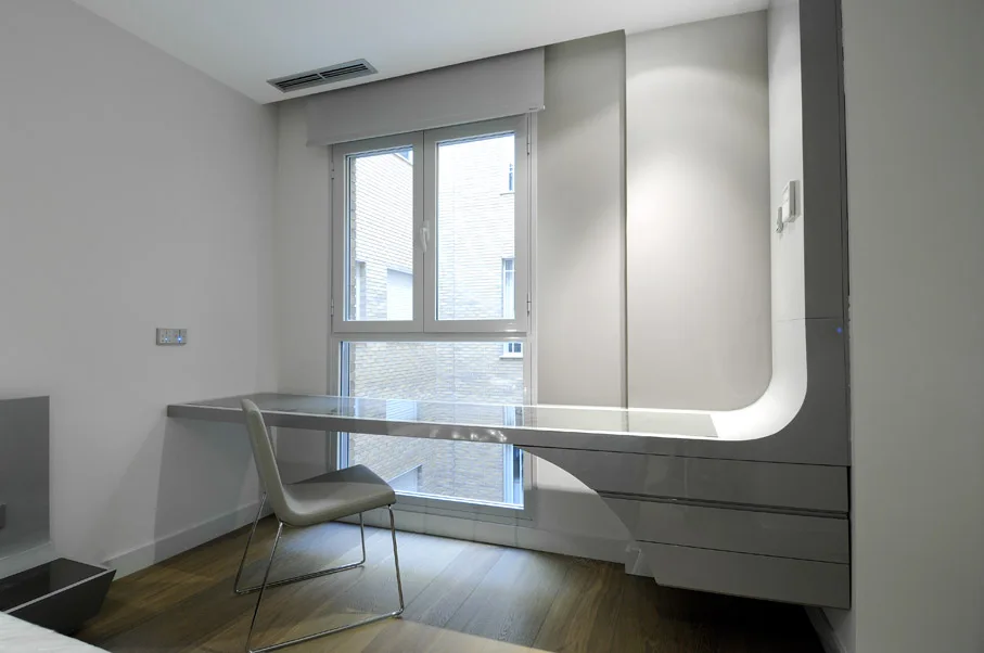 A minimalist room features a glossy gray cantilevered desk spanning a window, paired with a matching chair on wood floors.