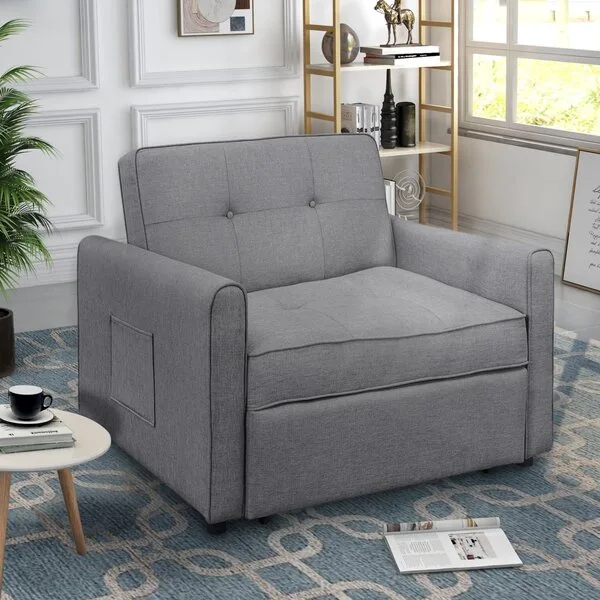 A grey fabric sleeper armchair with button tufting sits on a blue patterned rug next to a gold shelf and white paneled walls.
