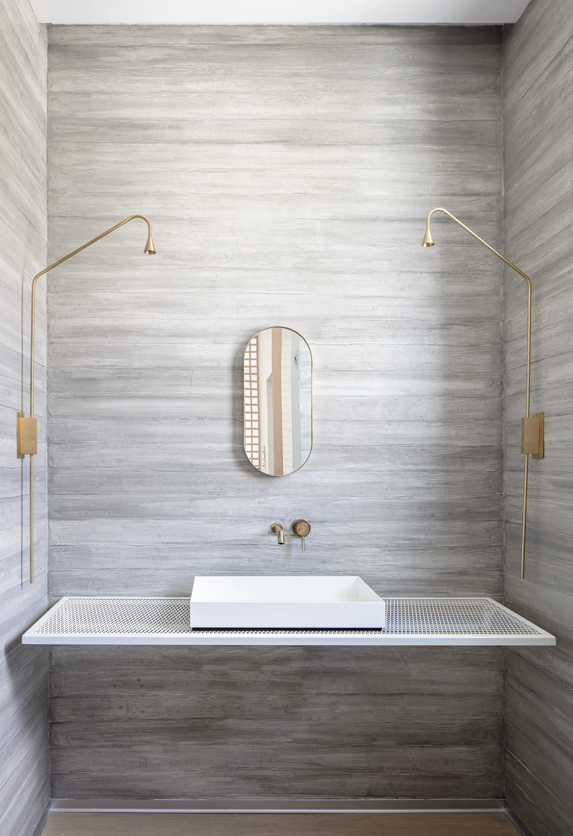 A minimalist bathroom vanity features a white mesh shelf holding a vessel sink against a gray textured wall with brass sconces and an oval mirror.