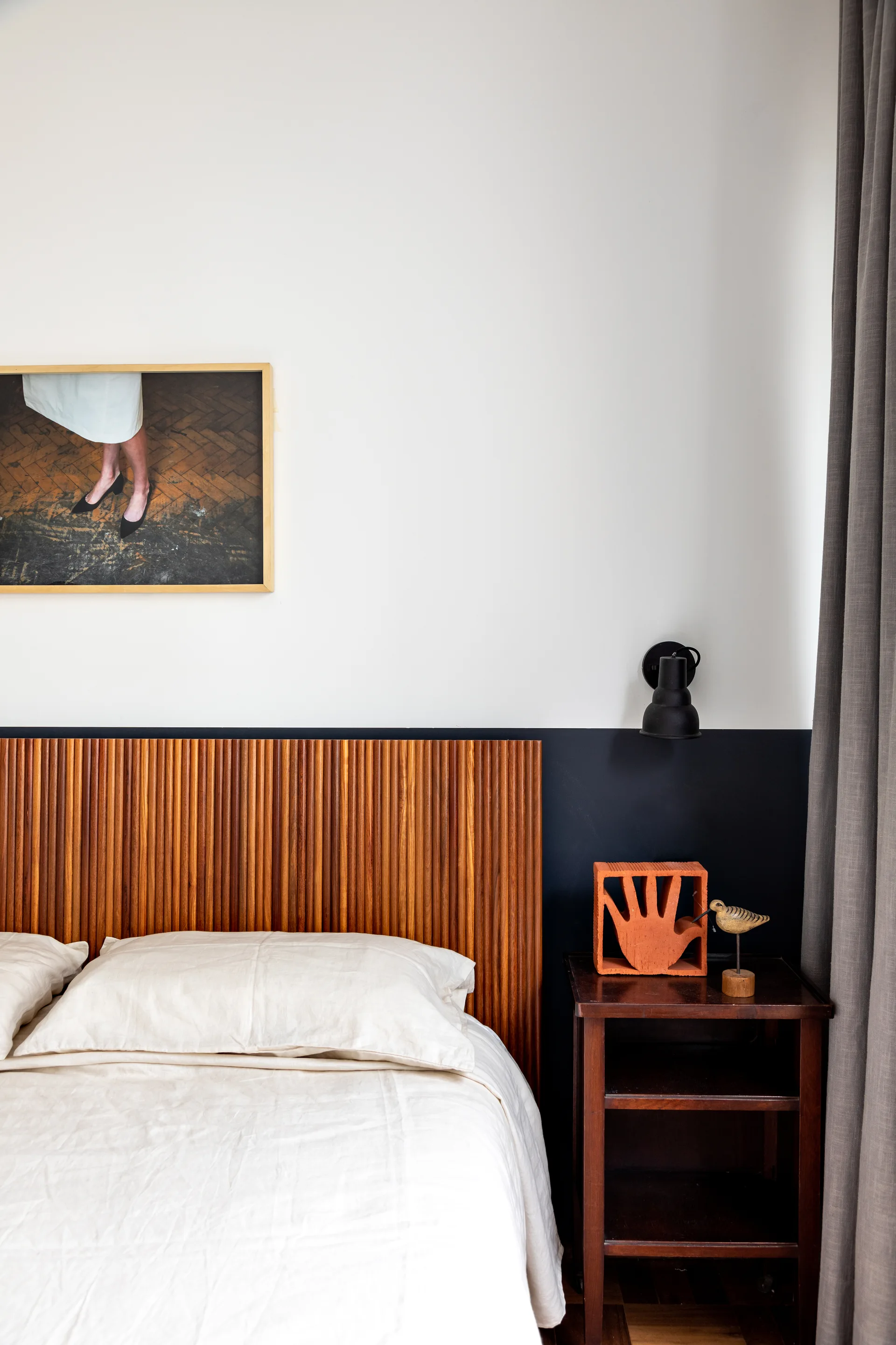 A bedroom corner featuring a fluted wood headboard against a color-blocked wall, a dark wood nightstand with terracotta decor, and a black wall sconce.