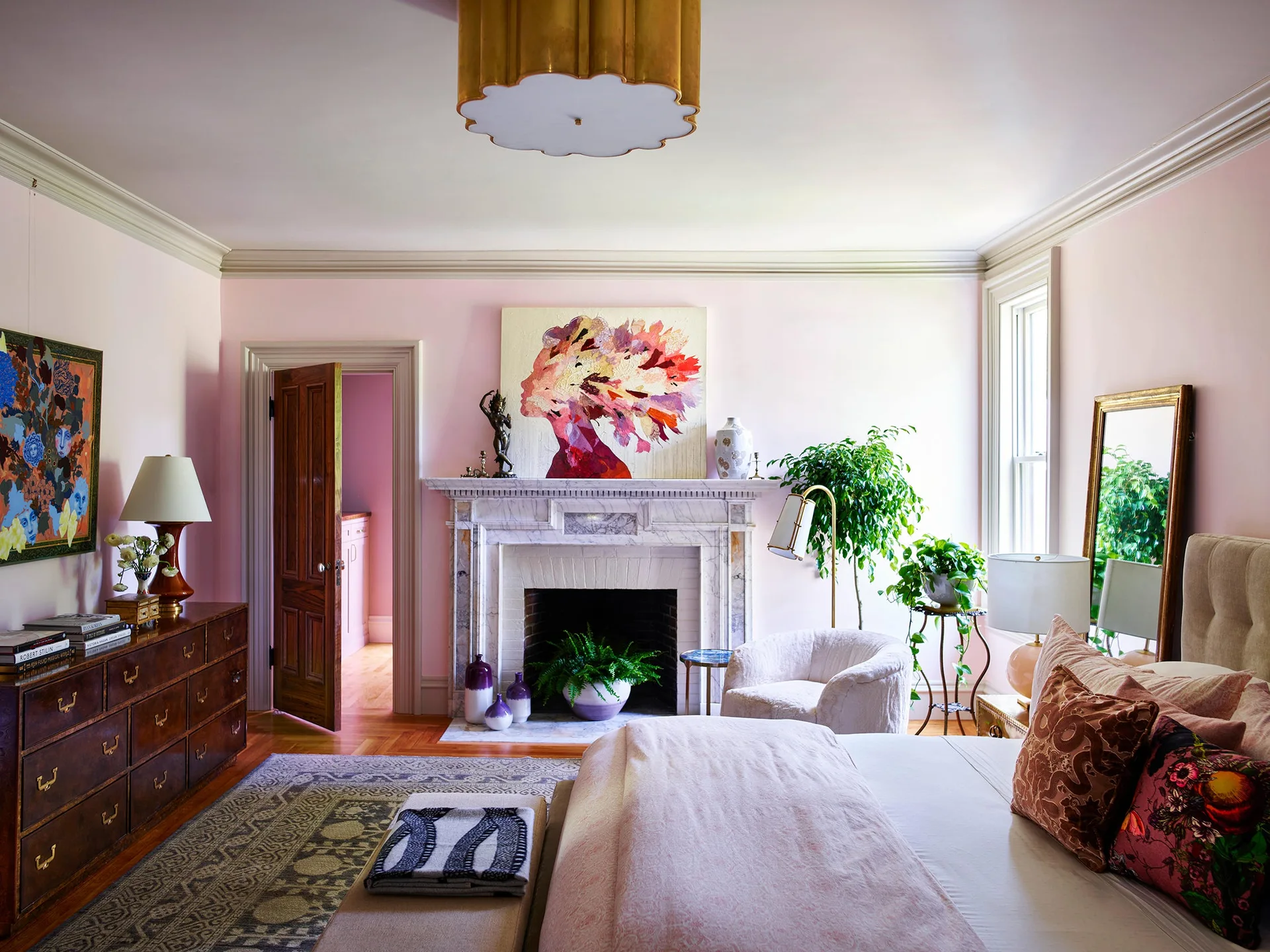 An eclectic bedroom features pale pink walls, a white marble fireplace with abstract art, a dark wood dresser, and a bed with textured pillows.