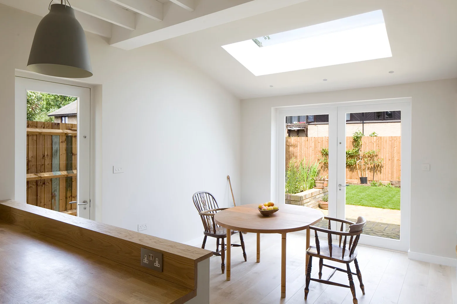 A sunlit dining room features a round wooden table, spindle chairs, and french doors opening to a green garden beneath a large skylight.