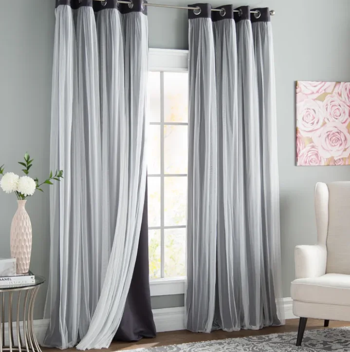 Layered grey and white sheer curtains hang over a window next to a white wingback armchair and a small side table.