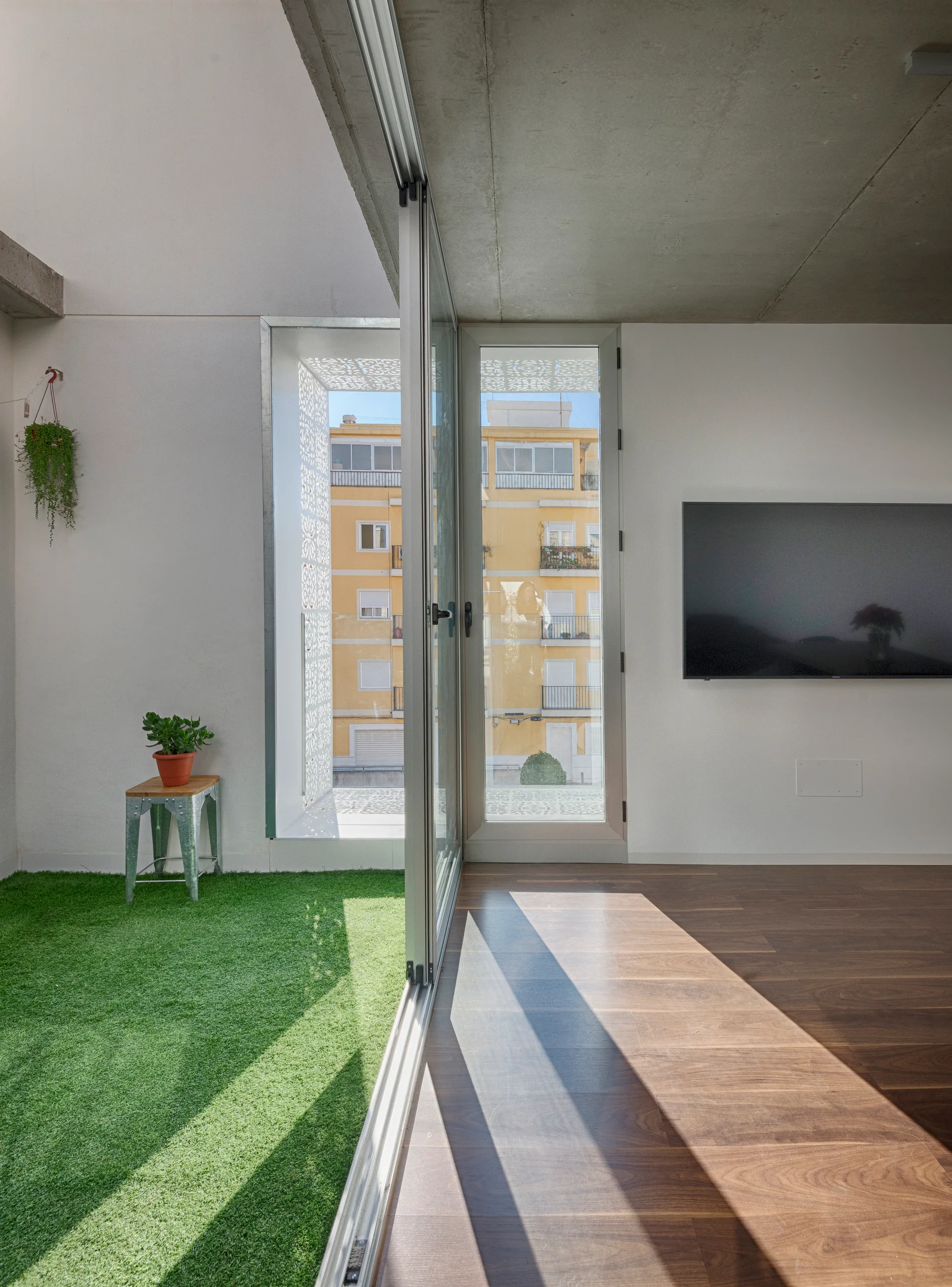 A modern living room with wood flooring and a wall-mounted TV opens via sliding glass doors to a balcony with artificial grass.