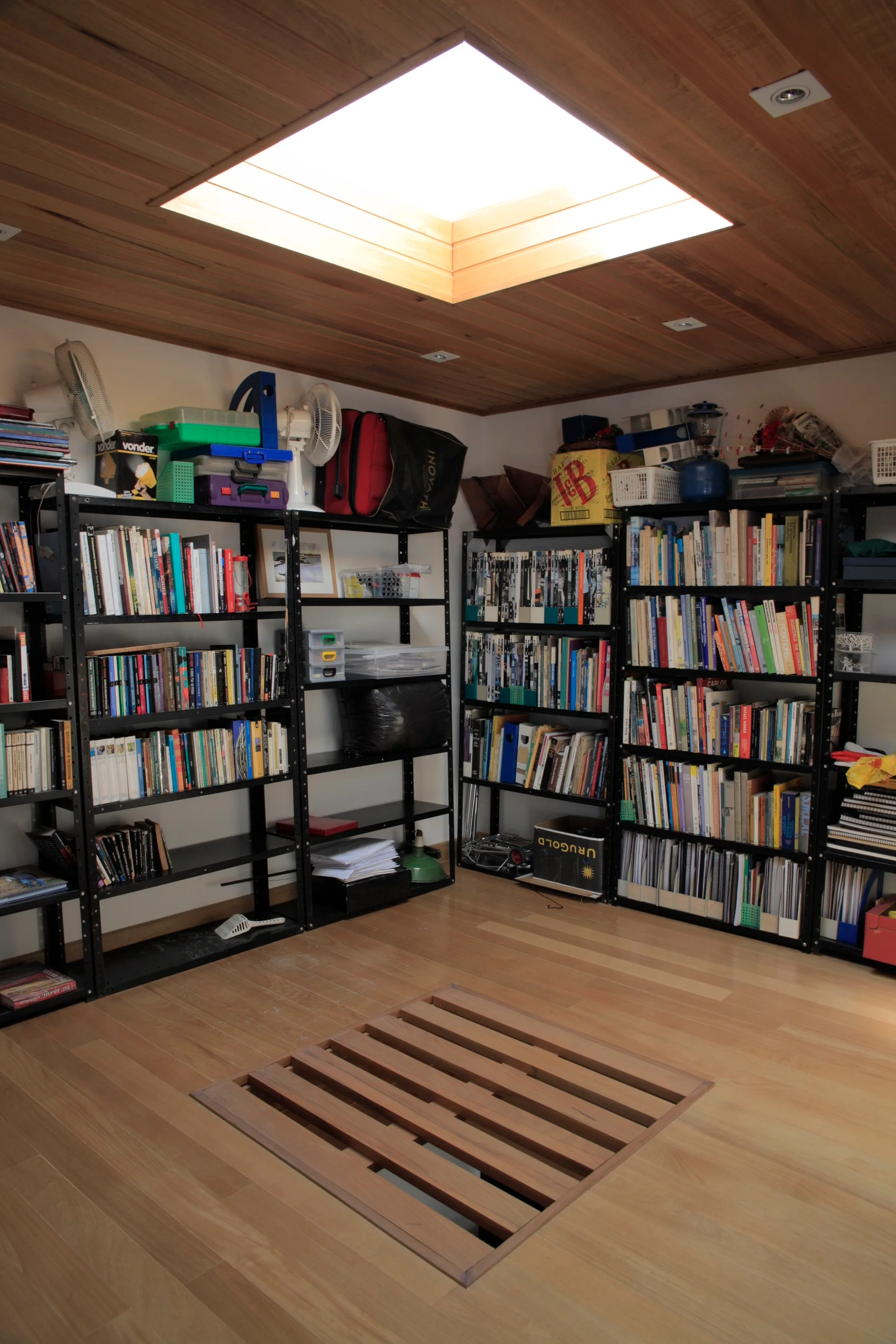 A home library filled with black metal bookshelves, featuring a wood plank ceiling with a skylight and a slatted wood hatch in the floor.