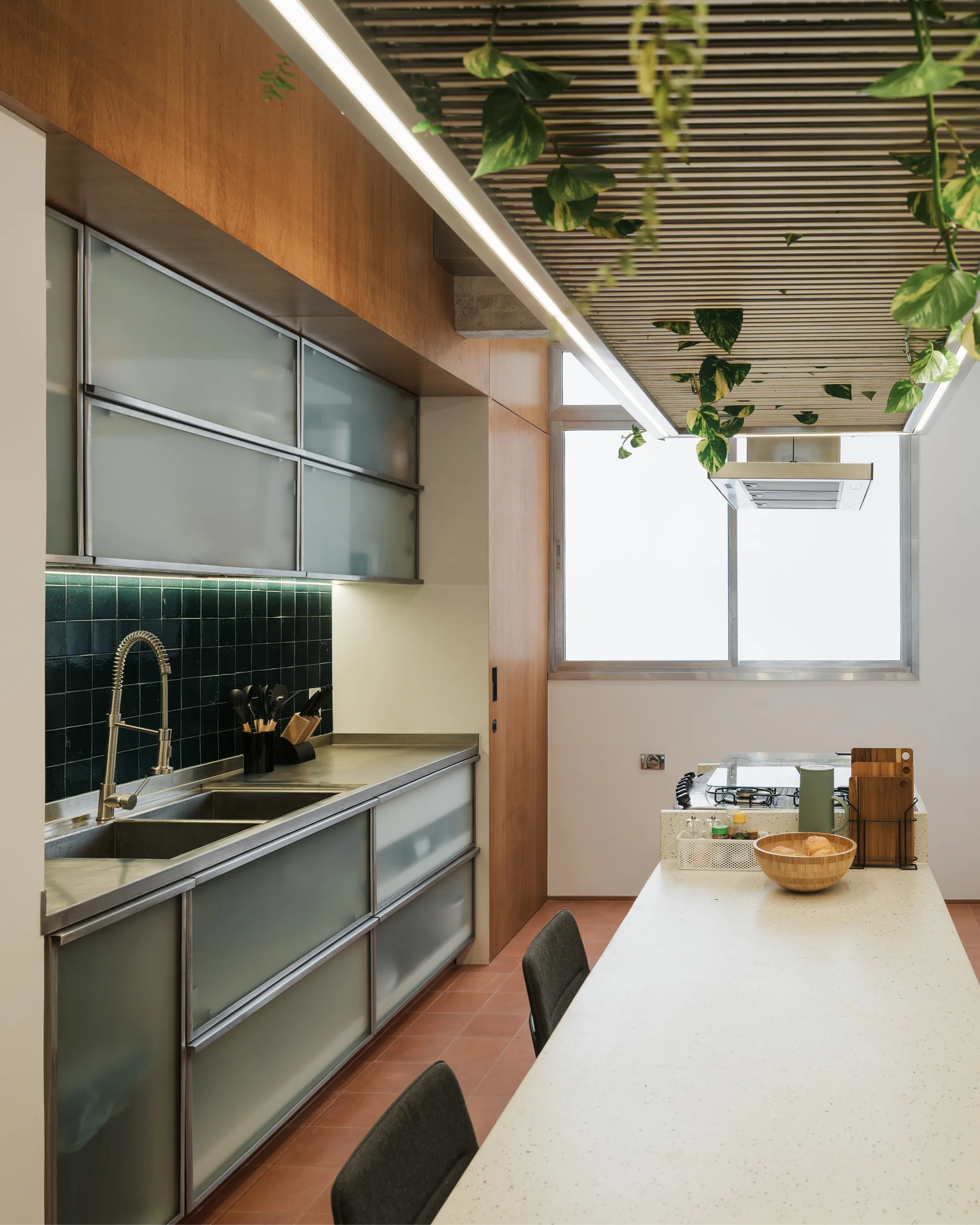 A modern kitchen featuring frosted glass cabinets, a dark green tiled backsplash, a long terrazzo island, and plants hanging from a slatted ceiling.
