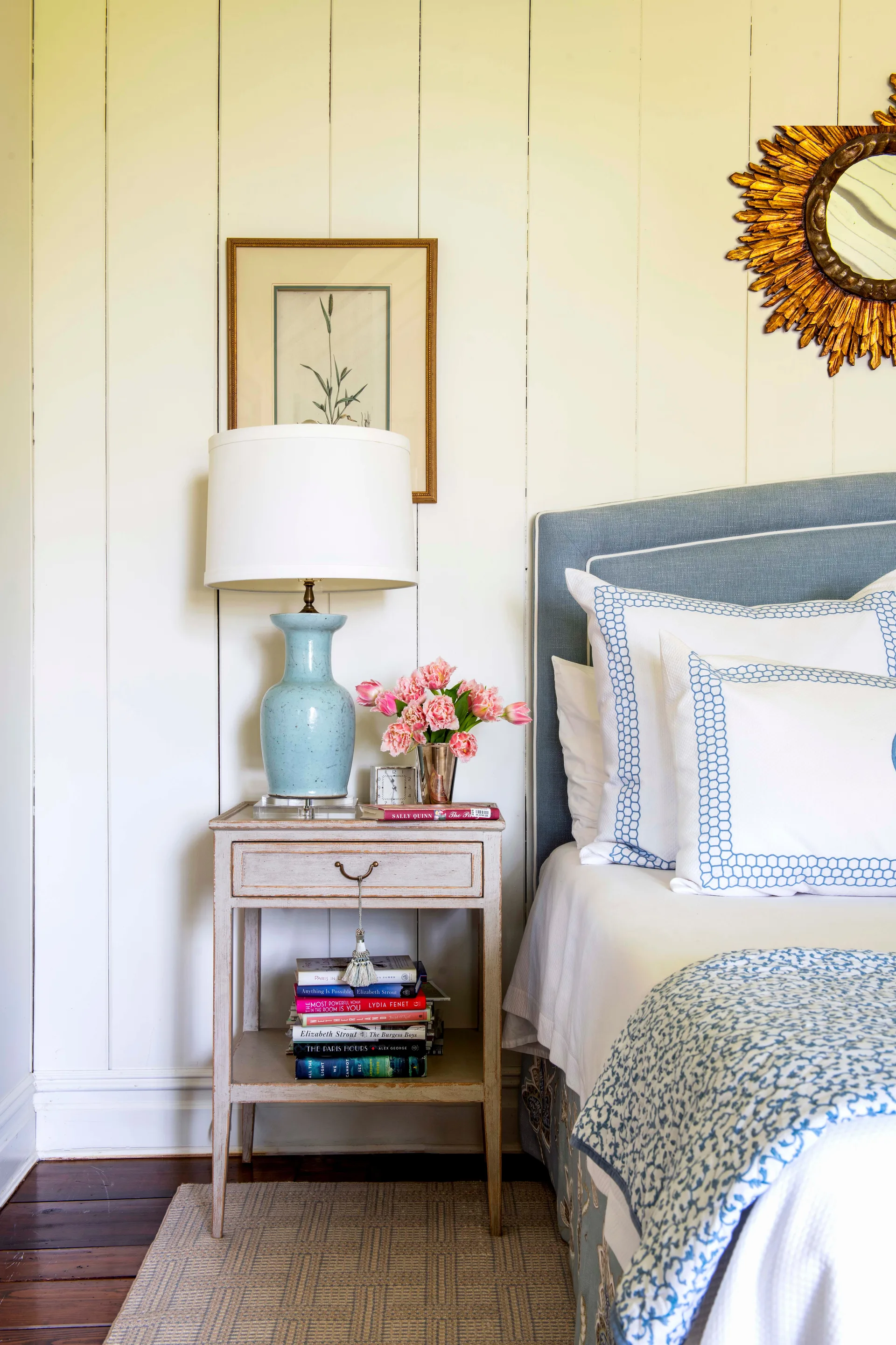 A cozy bedroom bedside vignette with a cream wood-paneled wall, a distressed wood nightstand, a blue ceramic lamp, and patterned blue bedding.
