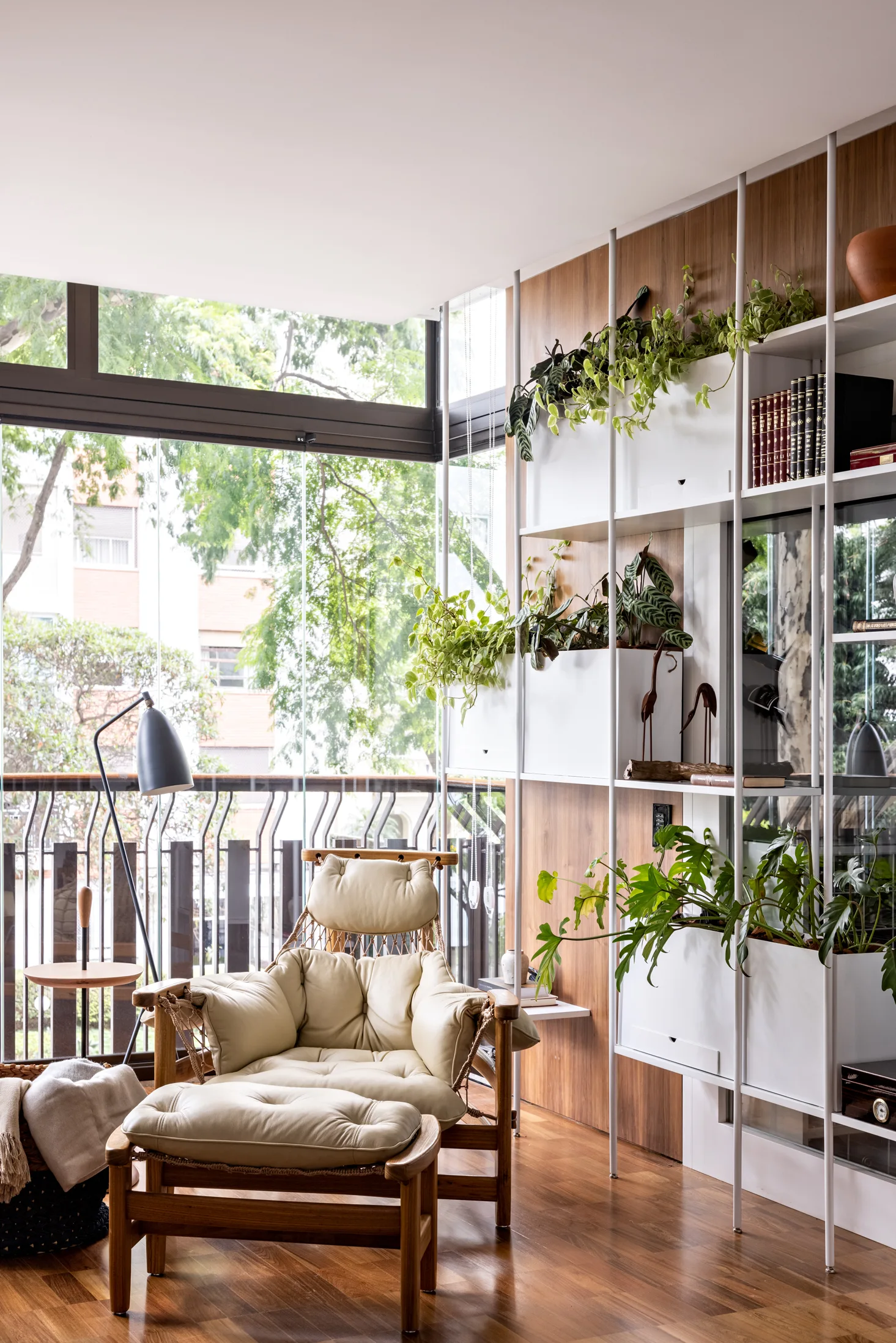 A beige leather lounge chair and ottoman sit next to a tall white shelving unit filled with houseplants in front of a glass wall.