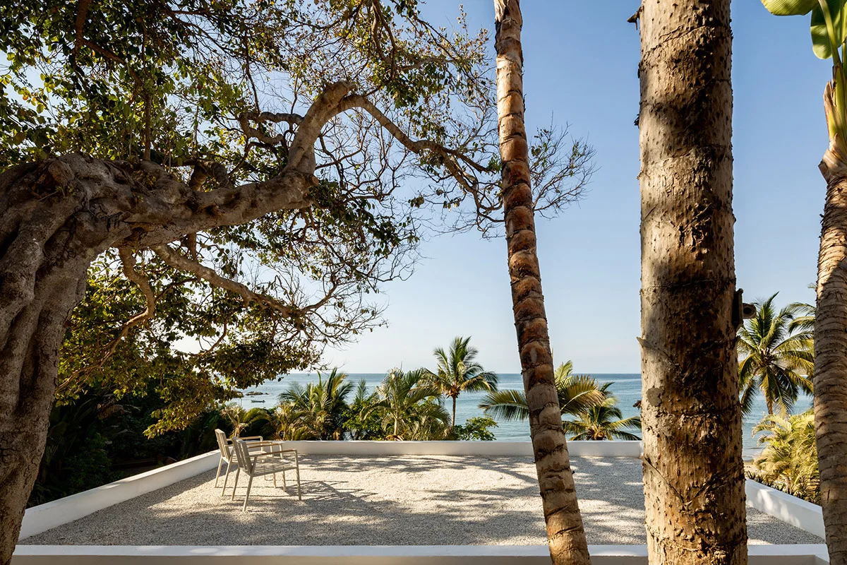 Two minimalist light grey chairs sit on a white gravel terrace overlooking the ocean framed by trees.