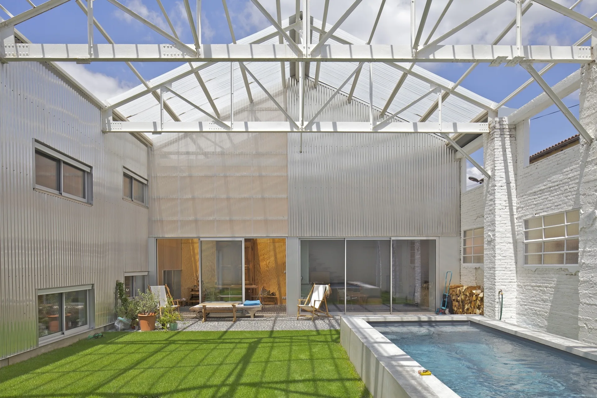 An outdoor courtyard with a concrete swimming pool, a grassy lawn, corrugated metal walls, and a white overhead metal truss structure.