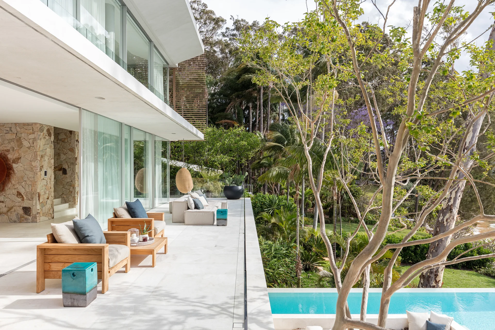 A modern white stone terrace features wood-framed lounge chairs with blue pillows next to a long swimming pool surrounded by lush trees.