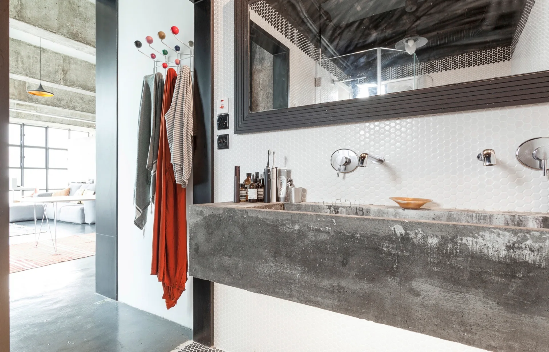 A bathroom vanity features a floating raw concrete trough sink set against a white hexagonal tiled wall below a large dark-framed mirror.