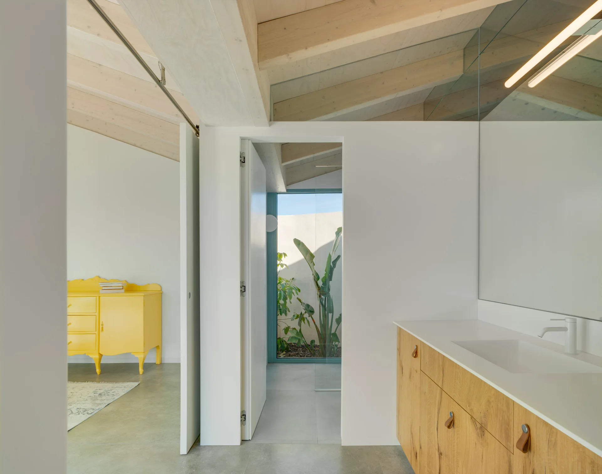 A modern bathroom features a light wood vanity with a white countertop under exposed ceiling beams, with a yellow dresser visible in the next room.