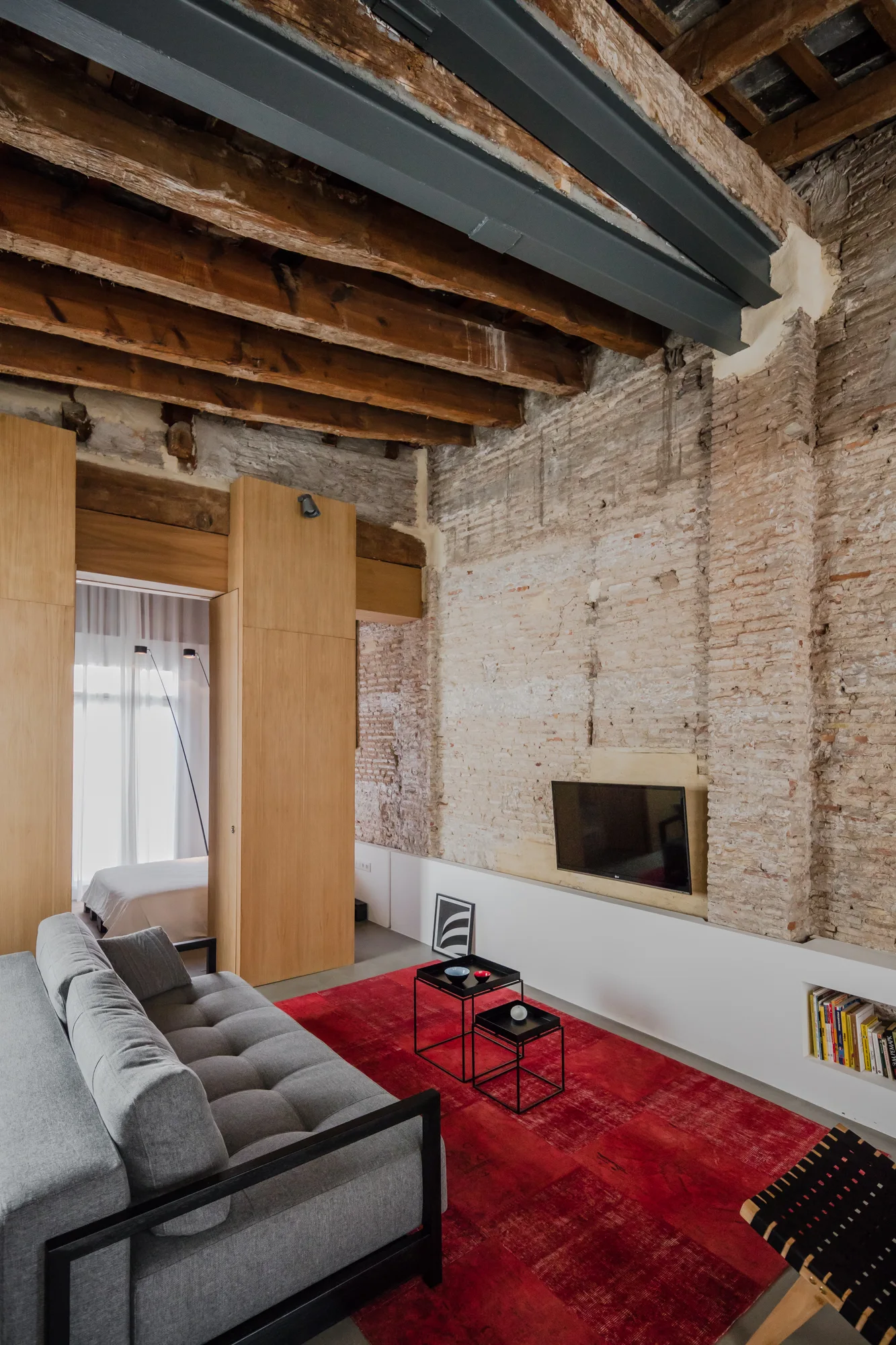 An industrial living room featuring an exposed brick wall, a grey tufted sofa on a red rug, wood ceiling beams, and oak wall panels.